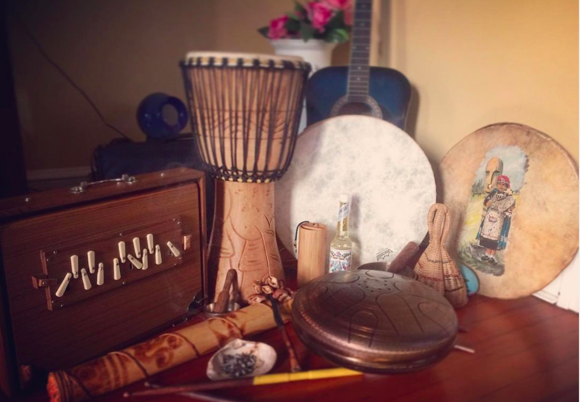 Collection of musical and decorative items including a wooden music box, a drum, a wooden-handled instrument, a hand drum, a guitar, a painted shield, a woven basket, and a bottle, arranged on a wooden surface.
