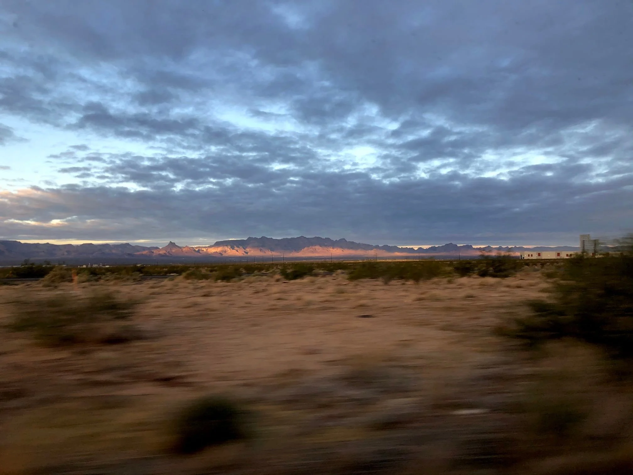 A desert landscape with mountains in the distance under a cloudy sky, possibly during sunrise or sunset.