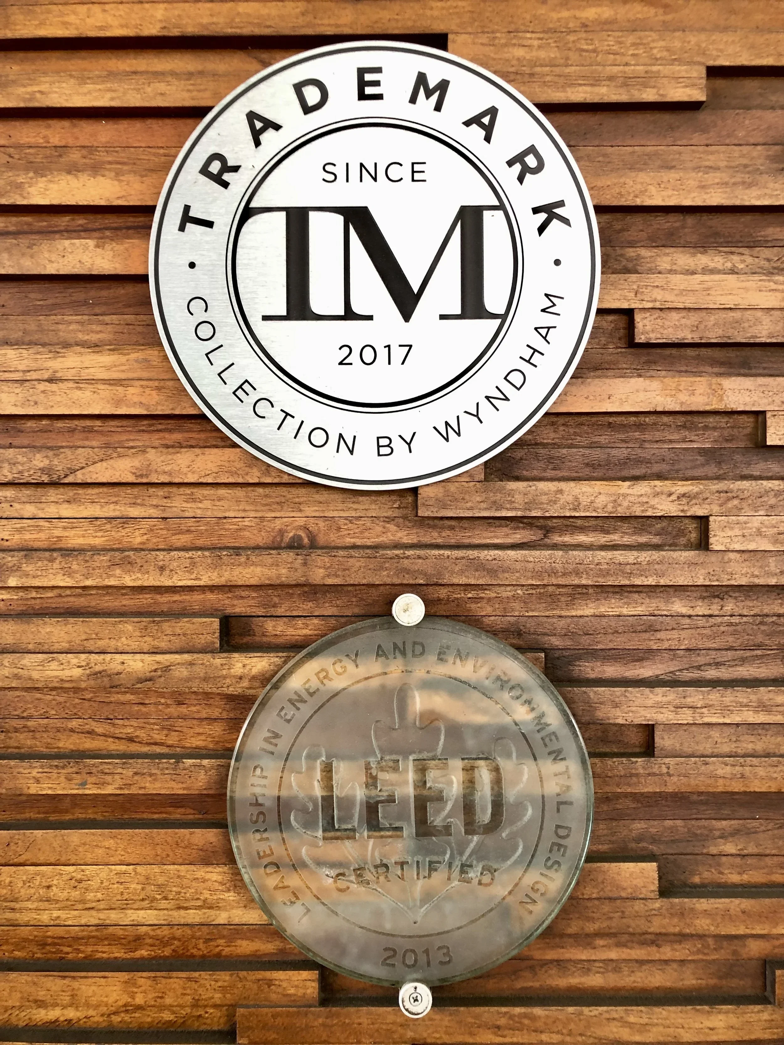 Wooden wall with a round metal sign that reads "TRADESMARK since 2017, collection by Wyndham." Below the sign, part of a transparent certificate with a maple leaf and the year 2013 is visible.