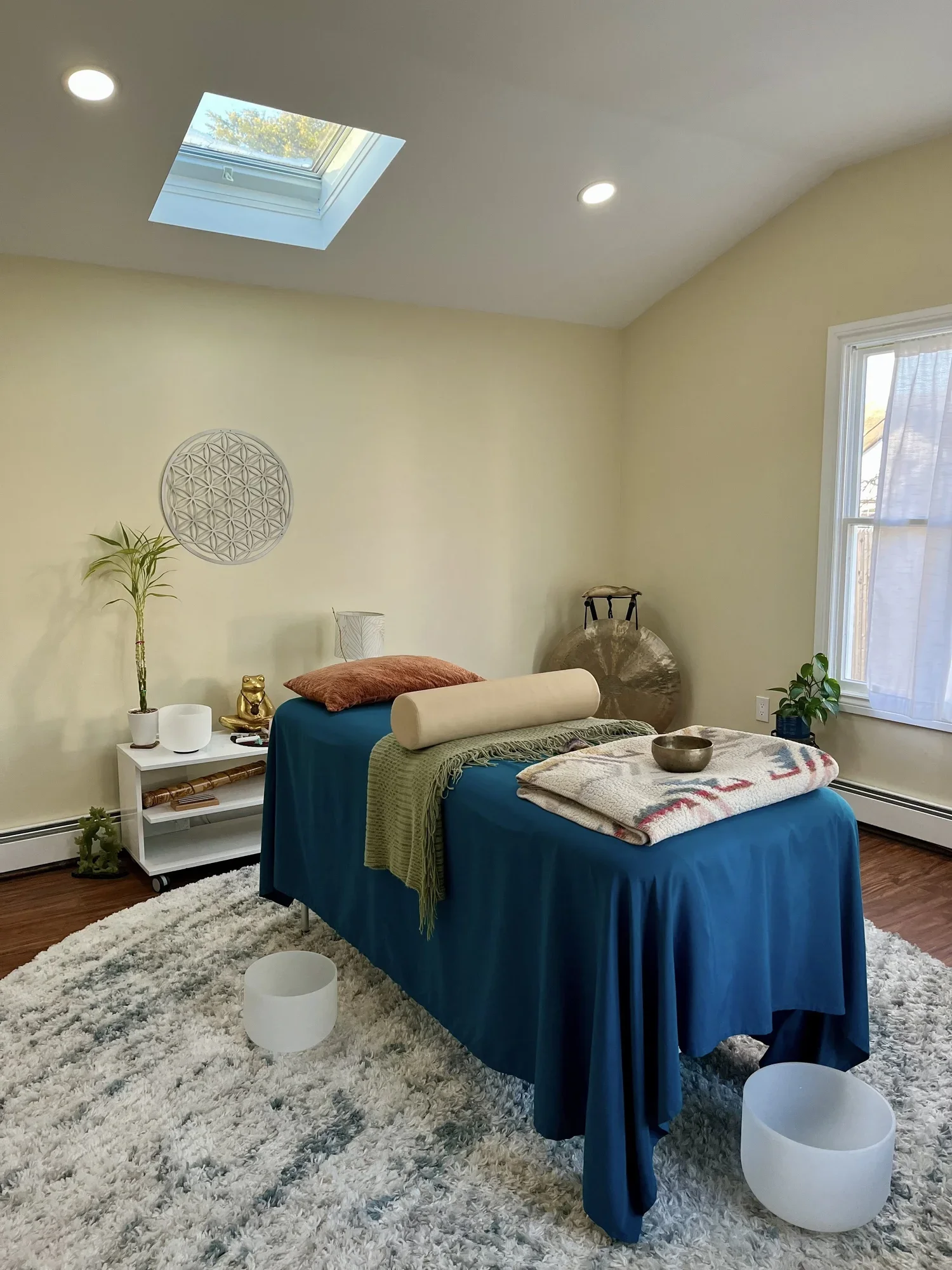 A cozy bedroom with a bed covered in a blue bedspread, a green throw, and a beige bolster pillow. There's a white shag rug on the floor, a gray and gold decorative object, a small white side table with plants and decor, and a window with sheer curtains. Two white cylindrical lamps sit on the floor, and the ceiling has a skylight and recessed lighting.