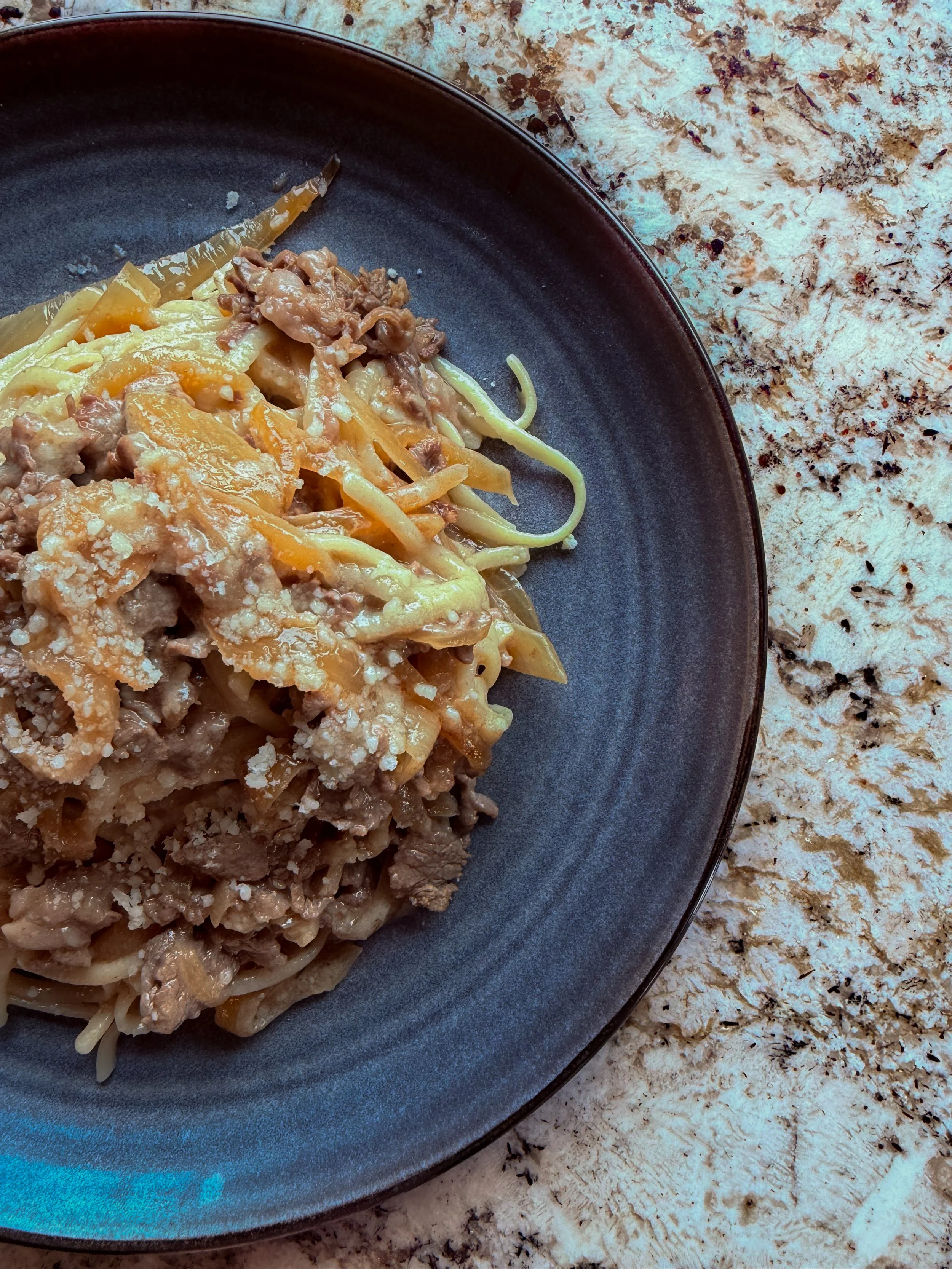 A plate of beef and vegetable stir-fry with noodles served on a dark plate, placed on a speckled white and brown marble countertop.