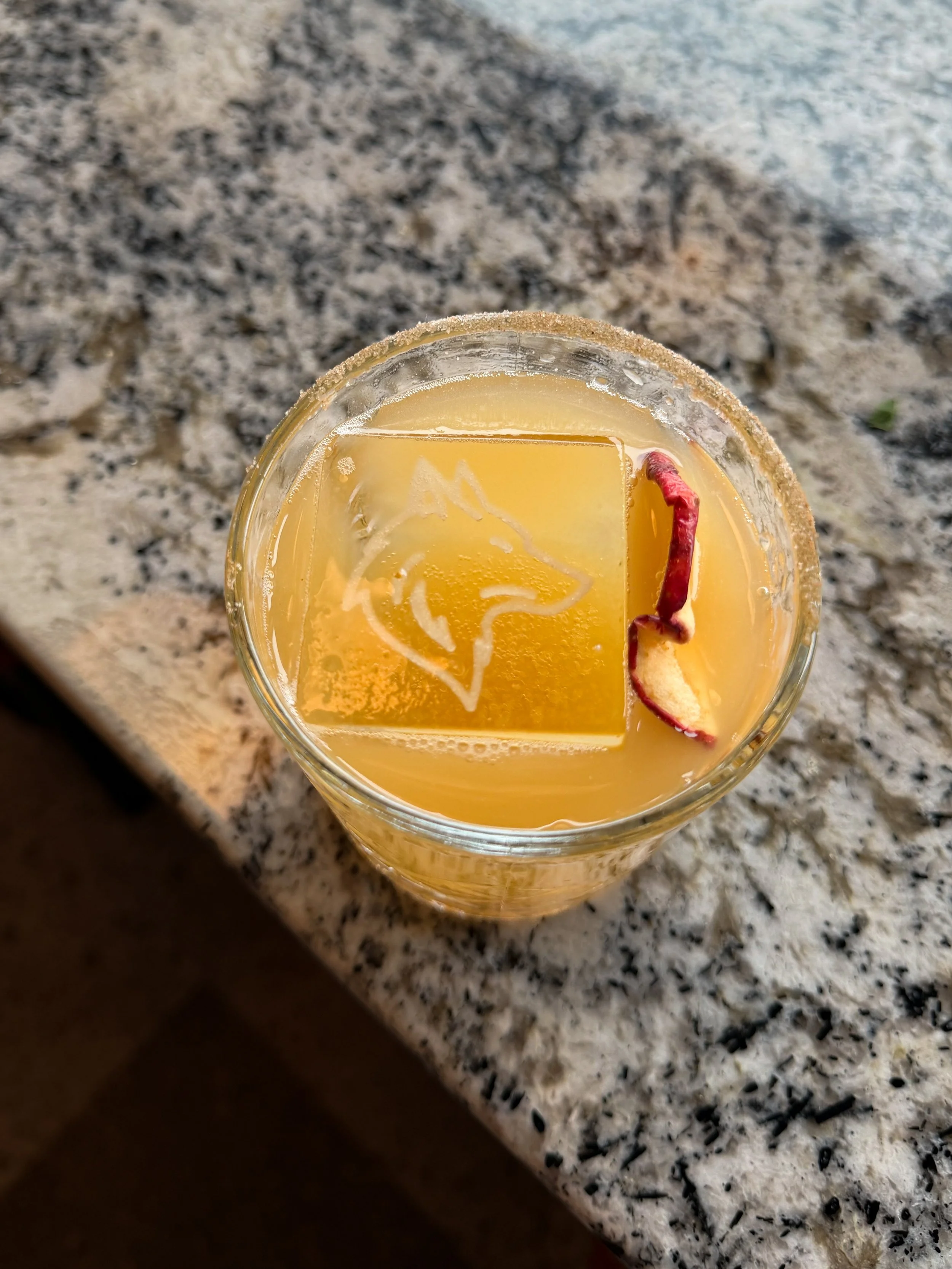 A glass of yellow cocktail with a large ice cube featuring a fox design, garnished with a rose petal and fruit peel, on a granite countertop.