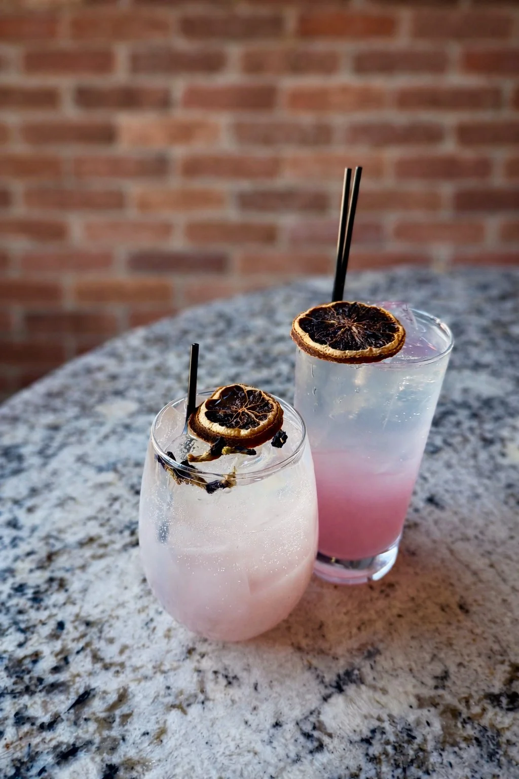 Two cocktails with dried lemon slices garnishes and black straws on a granite table with a brick wall background.