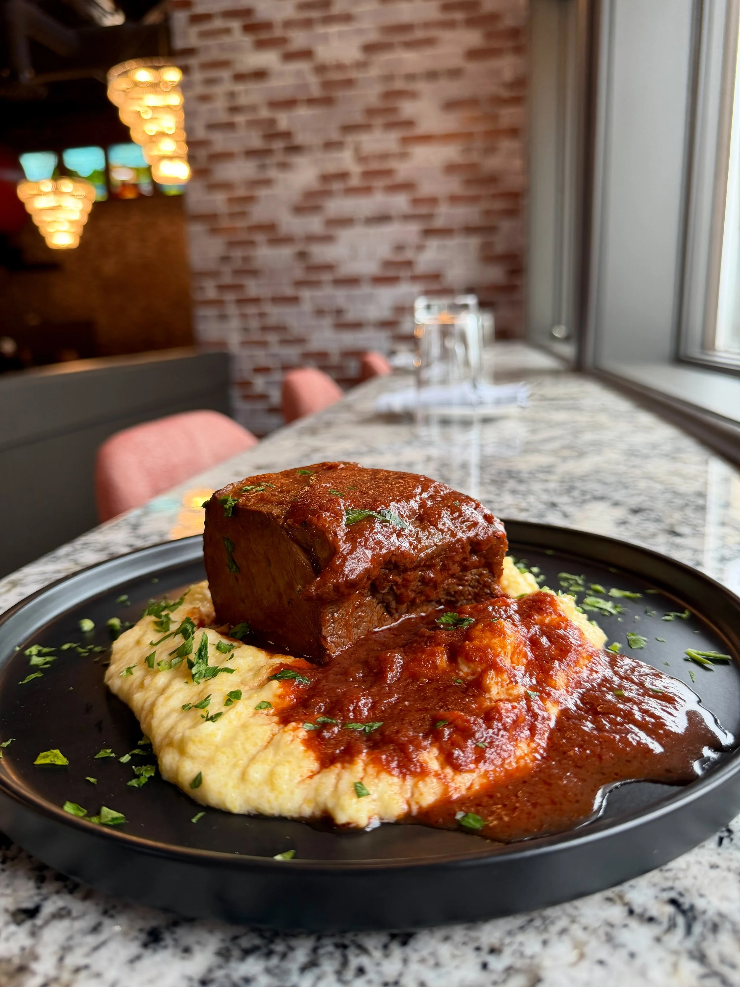 Plate of braised beef with mashed potatoes and gravy in a restaurant with a window and brick wall in the background.