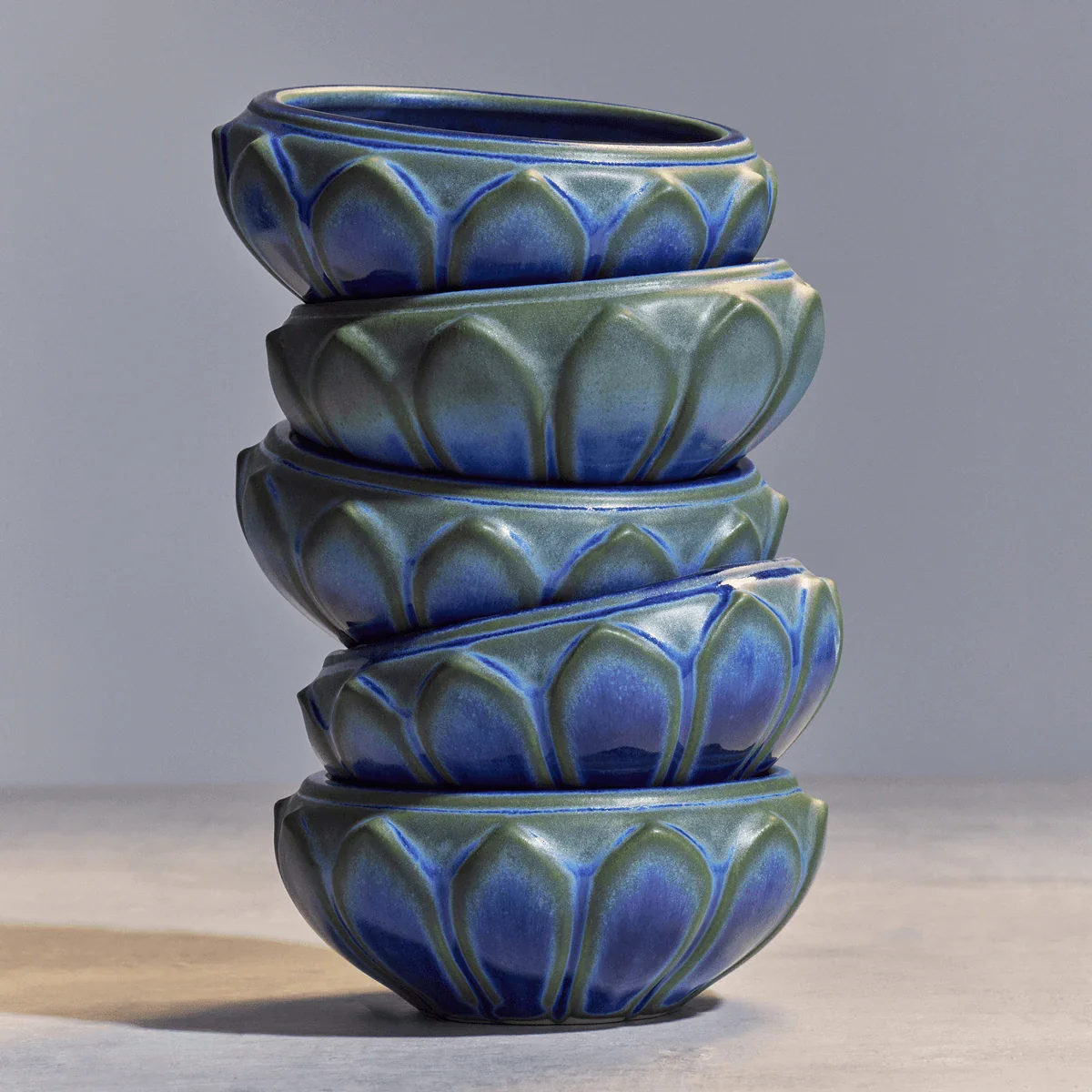 Stack of five ceramic bowls with a blue and green glaze, each with a decorative petal pattern, placed on a light wooden surface.