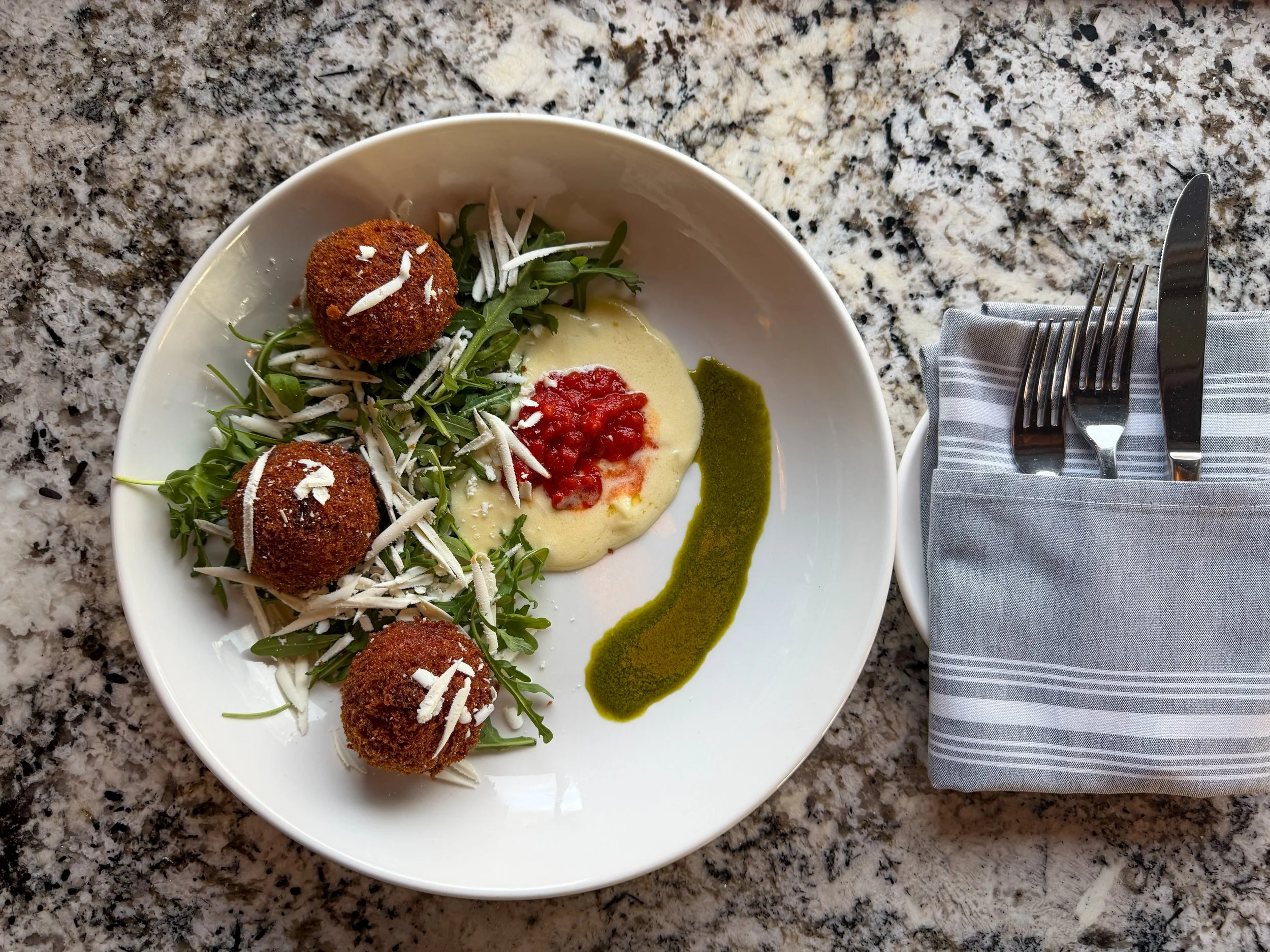 Plate with three breaded and fried round appetizers on a bed of arugula and shredded cheese, accompanied by a dollop of yellow sauce with red topping and a green sauce on the side, with utensils wrapped in a gray napkin on a granite countertop.