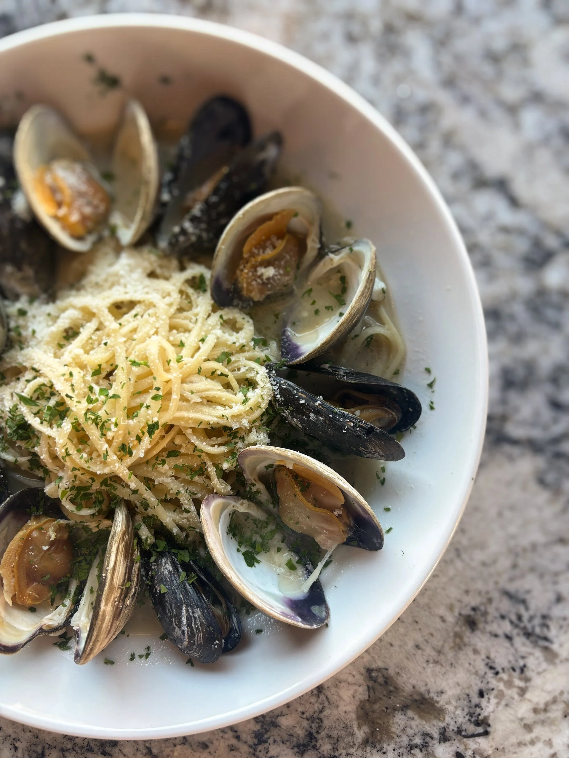 Bowl of spaghetti with mussels in cream sauce, topped with grated cheese and parsley, on a granite countertop.