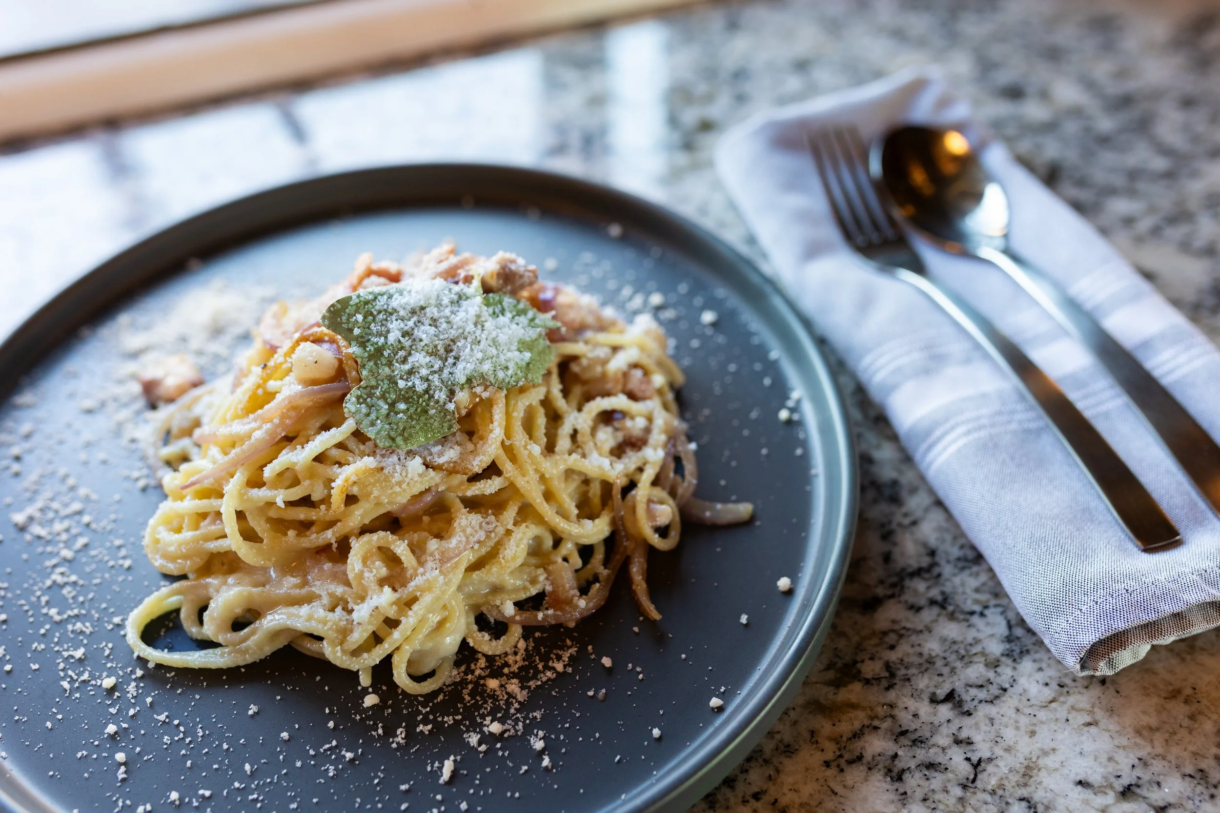 Plate of spaghetti pasta with grated cheese and a basil leaf on top, next to a fork and spoon on a napkin, on a granite countertop.