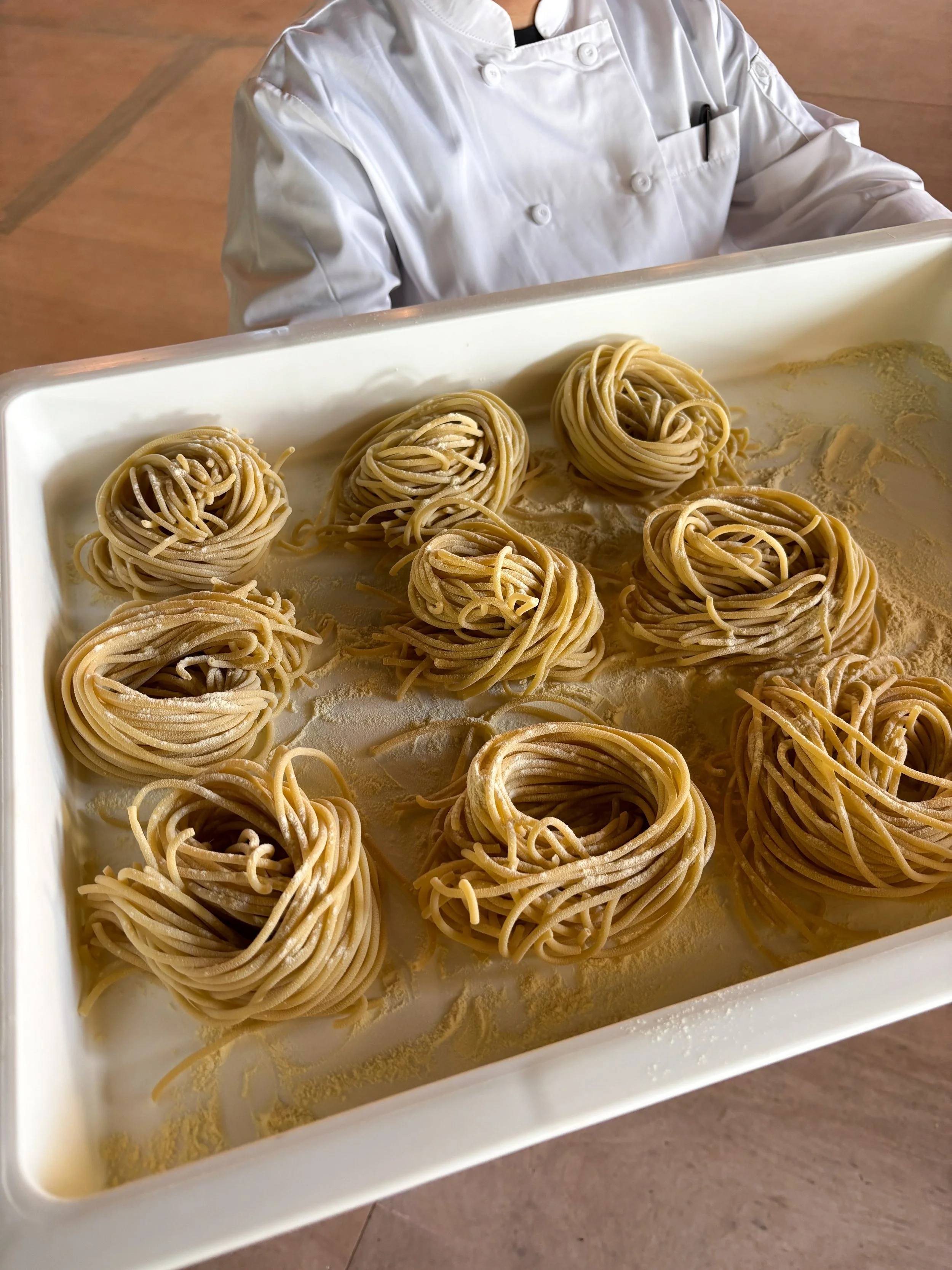 Chef in white uniform holding a tray of freshly made, uncooked spaghetti nests coated in flour.