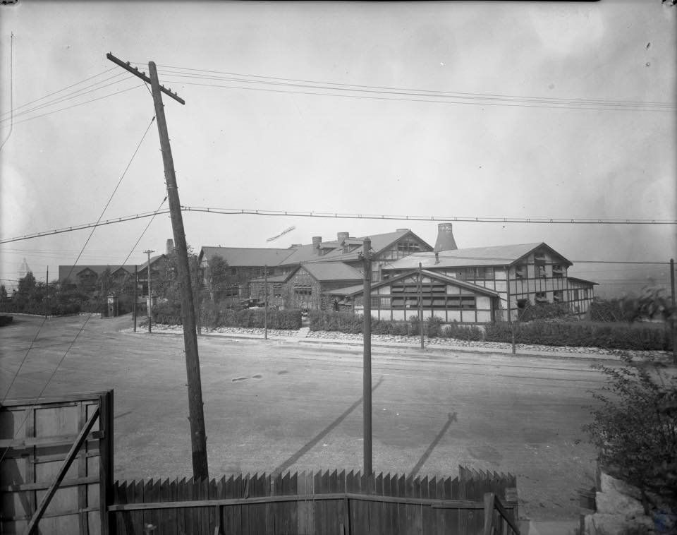 Black and white photograph of an old train station or railway building with multiple tracks and a water tower in the background, viewed from a fenced area with power lines and utility poles in the foreground.