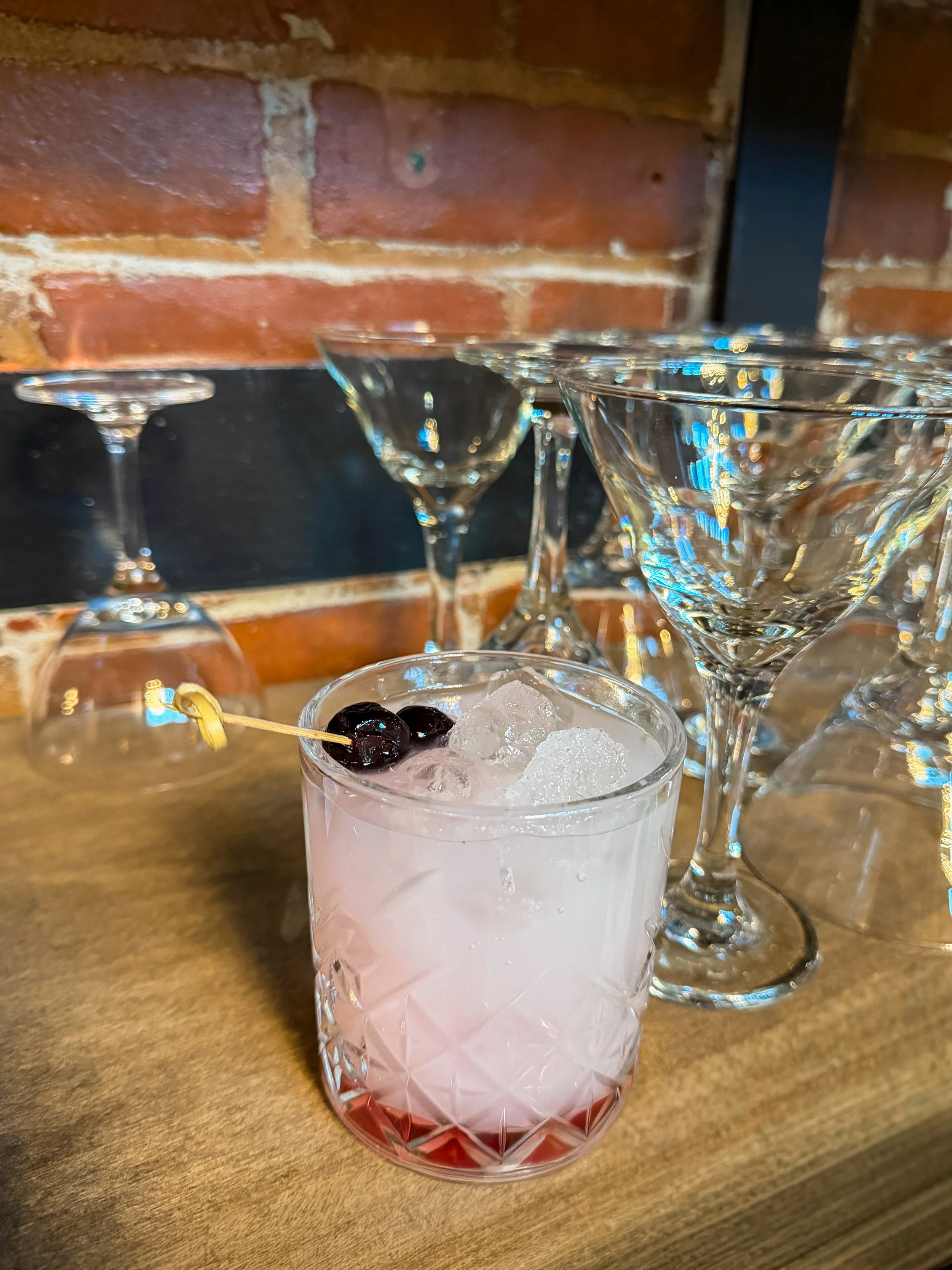 A cocktail glass filled with ice and a pink-colored drink, garnished with two black cherries on a toothpick, set on a wooden surface with empty martini glasses in the background and a brick wall behind.
