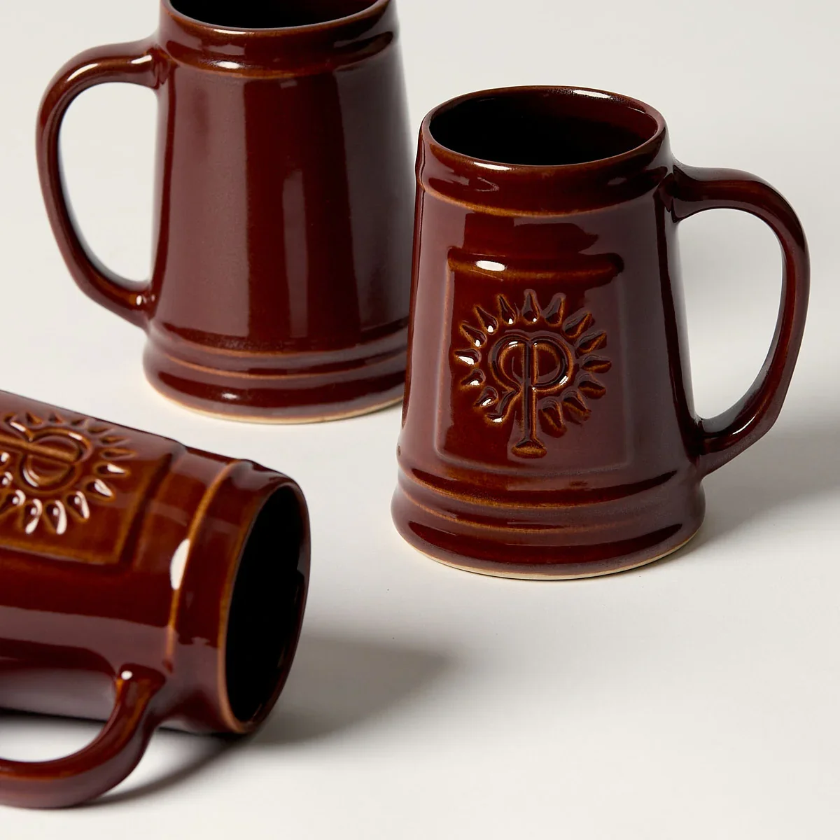 Three brown ceramic coffee mugs, two upright and one lying on its side, with a raised symbol of a chi-rho inside a sunburst pattern.