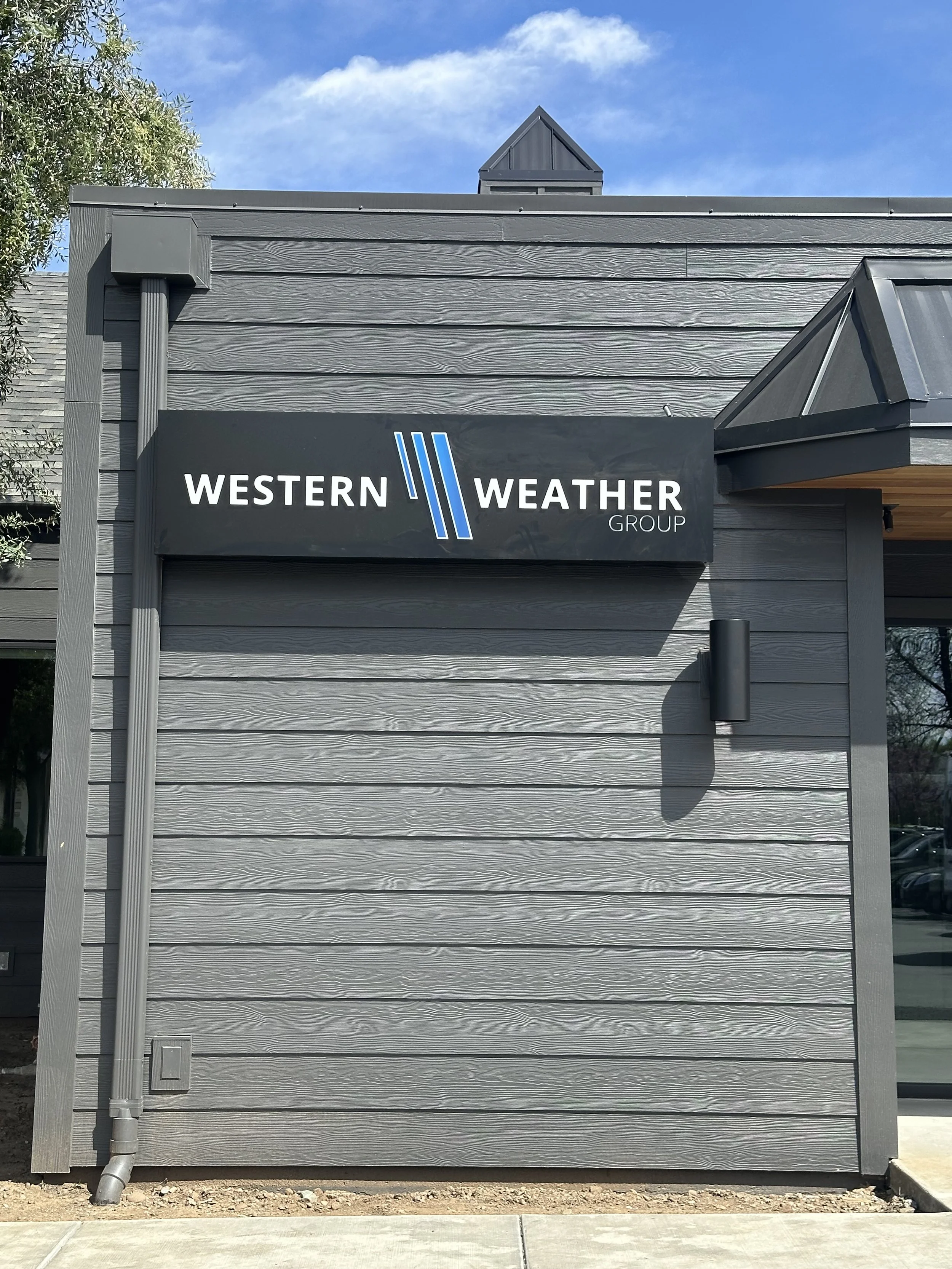 Sign for Western Weather Group on the exterior of a gray building with modern siding, against a blue sky with some clouds.