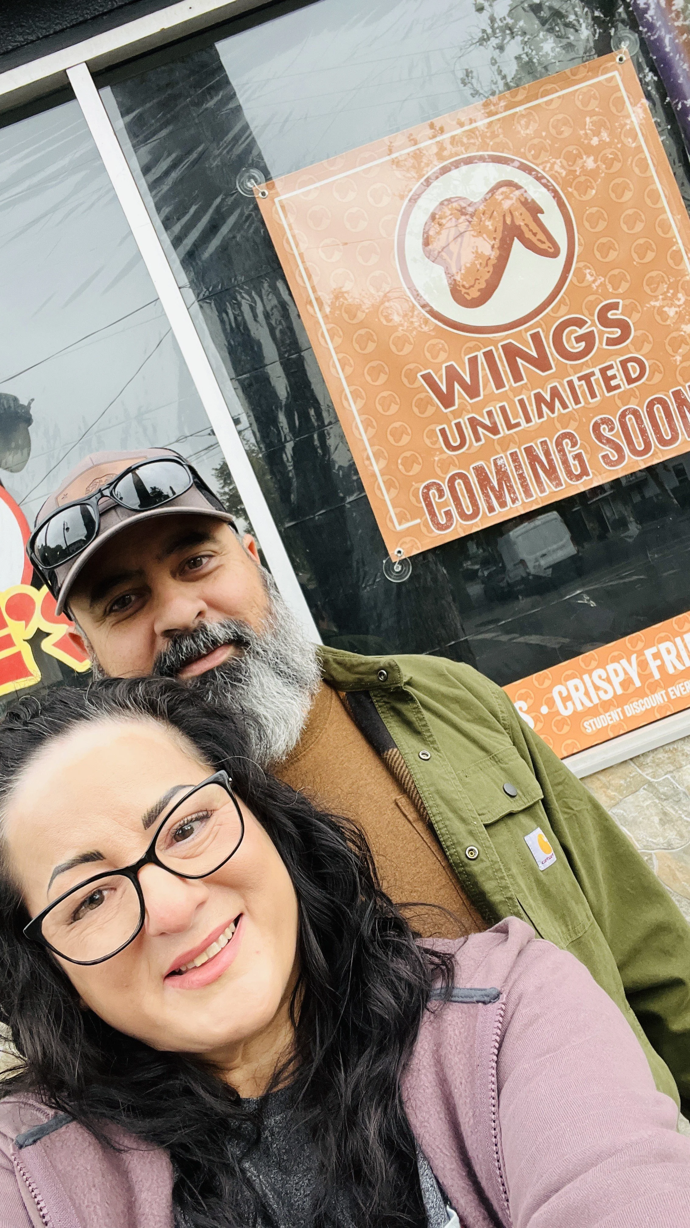 A smiling woman with black curly hair and glasses takes a selfie with a bearded man with gray hair and a baseball cap outside a restaurant. The restaurant sign indicates that wings are coming soon and are unlimited, with a placeholder for crispy fried wings.