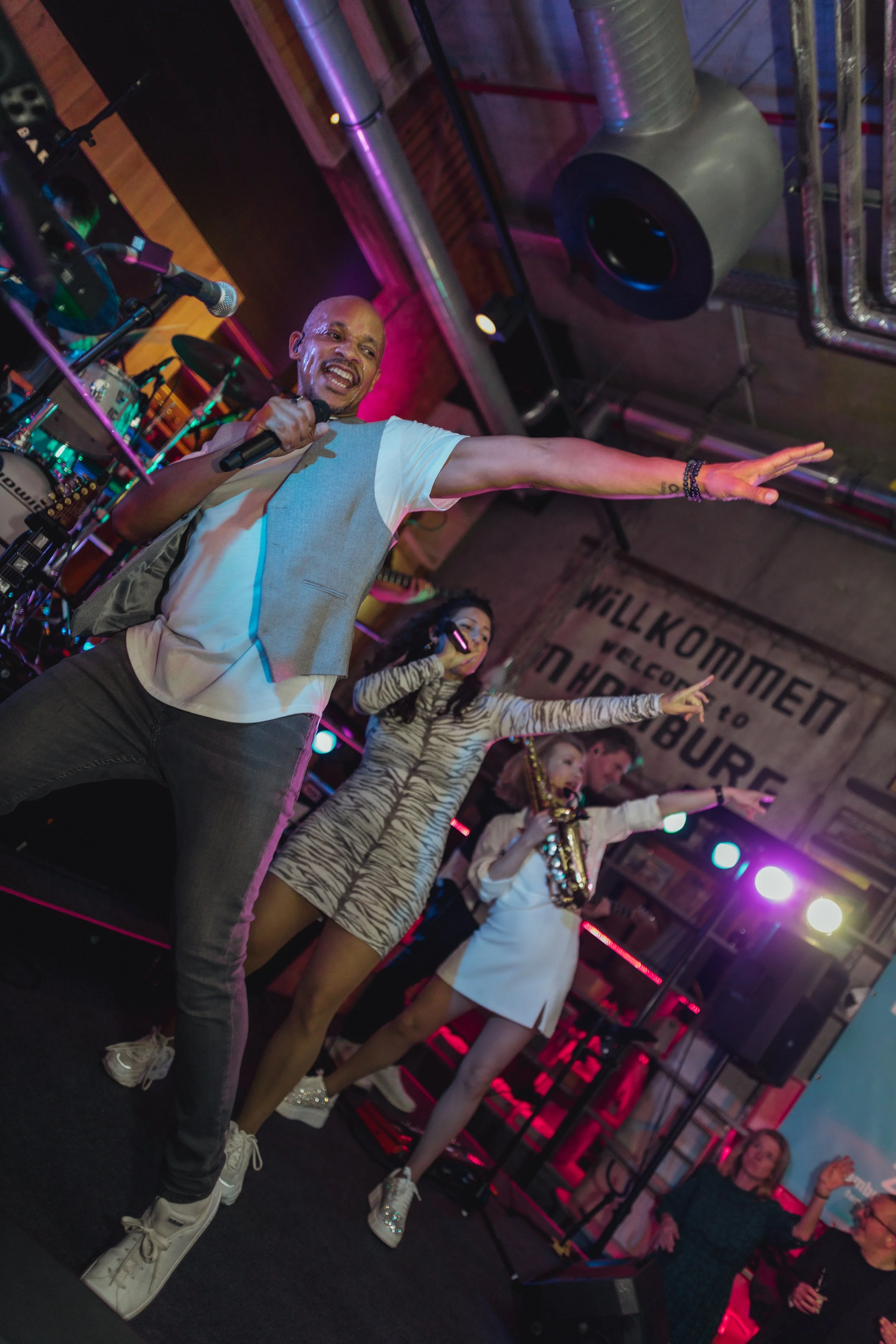 Mehrere Musiker und Sänger performen in einer Bar oder einem Club mit Neon-beleuchteter Bühne, inklusive einem Mann mit Mikrofon, einer Frau mit Mikrofon, einer Frau mit Saxophon, und mehreren Publikum im Hintergrund.