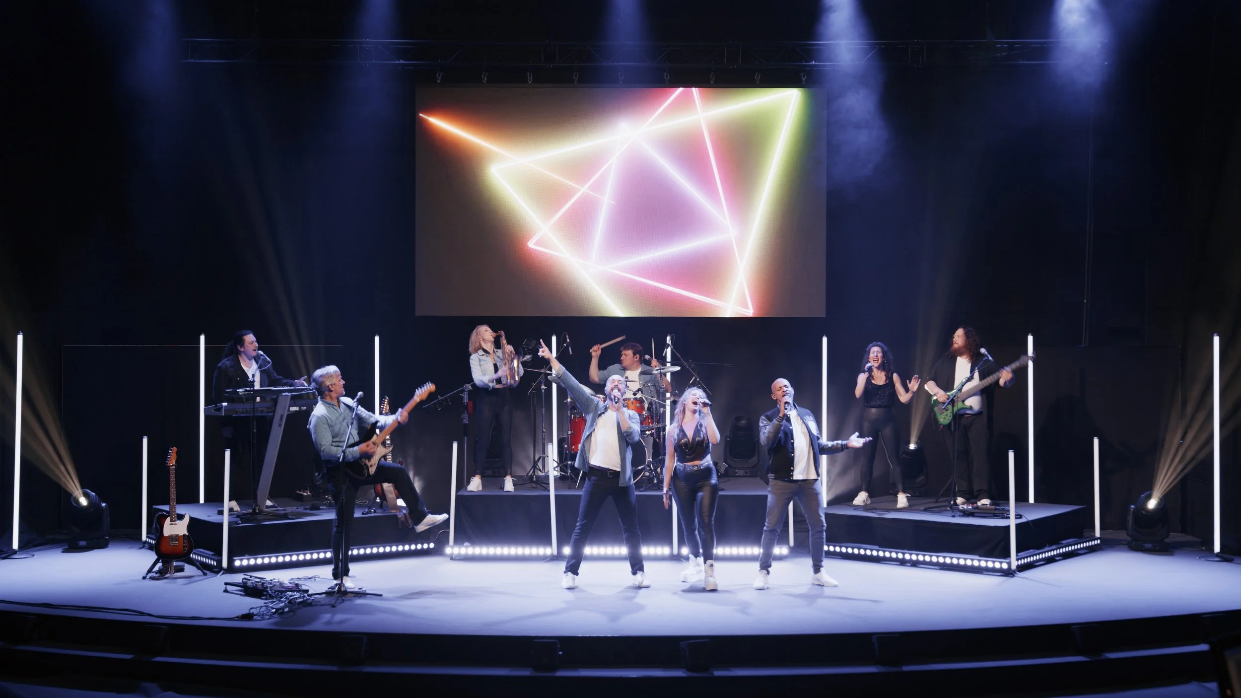 Eine Band mit mehreren Mitgliedern, die auf einer Bühne bei einem Live-Konzert auftreten. Es gibt Sänger, Gitarristen, einen Keyboardspieler, einen Drummer und einen Bassisten. Die Bühne ist mit Lichtstangen und LED-Leuchten dekoriert, im Hintergrund läuft eine bunte Lichtshow auf einem großen Bildschirm.