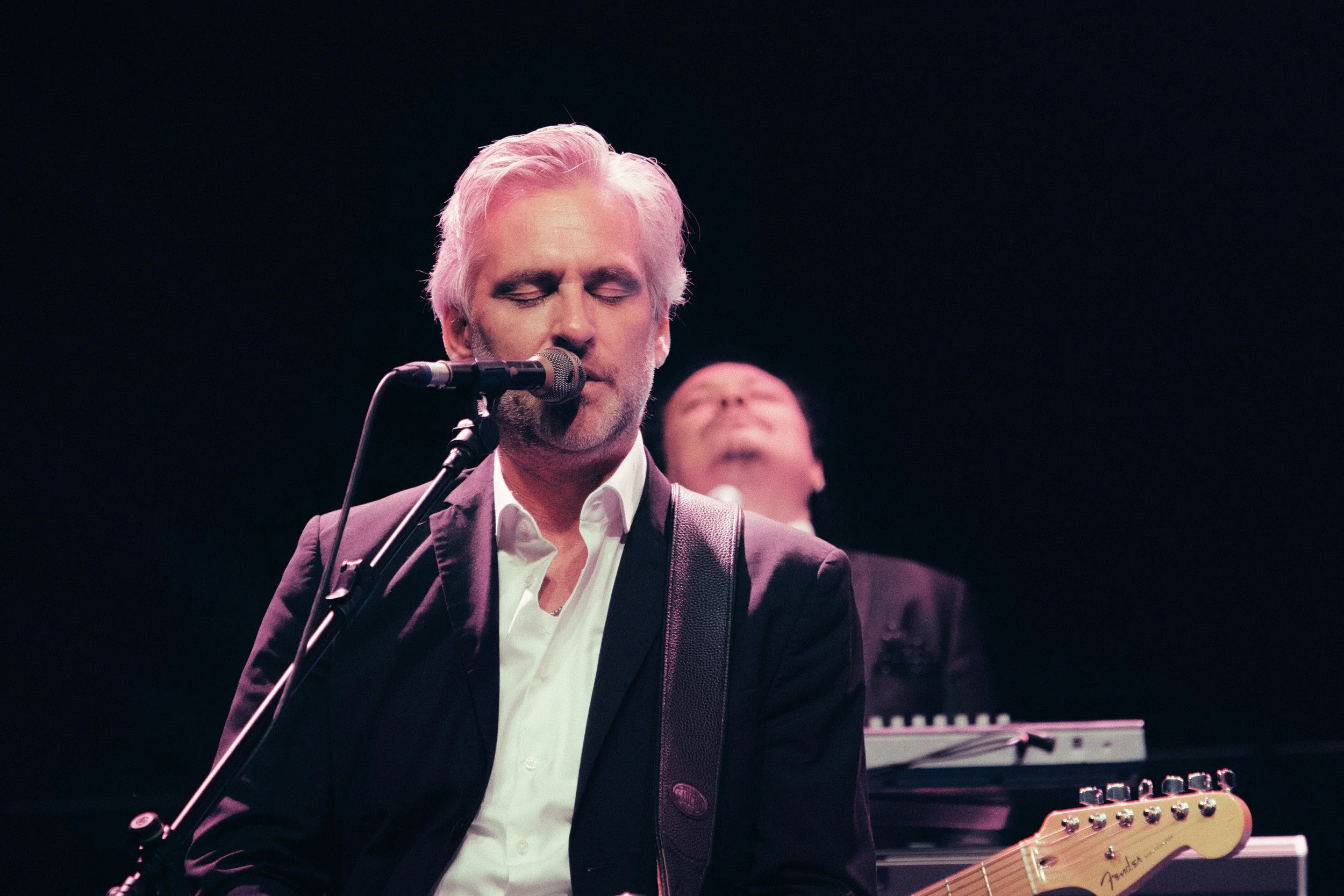 Ein Mann mit grauem Haaren und weißem Hemd, der Gitarre spielt und singt, im Hintergrund ist eine Person mit geschlossenen Augen und einem Keyboard zu sehen, bei einem Live-Konzert.