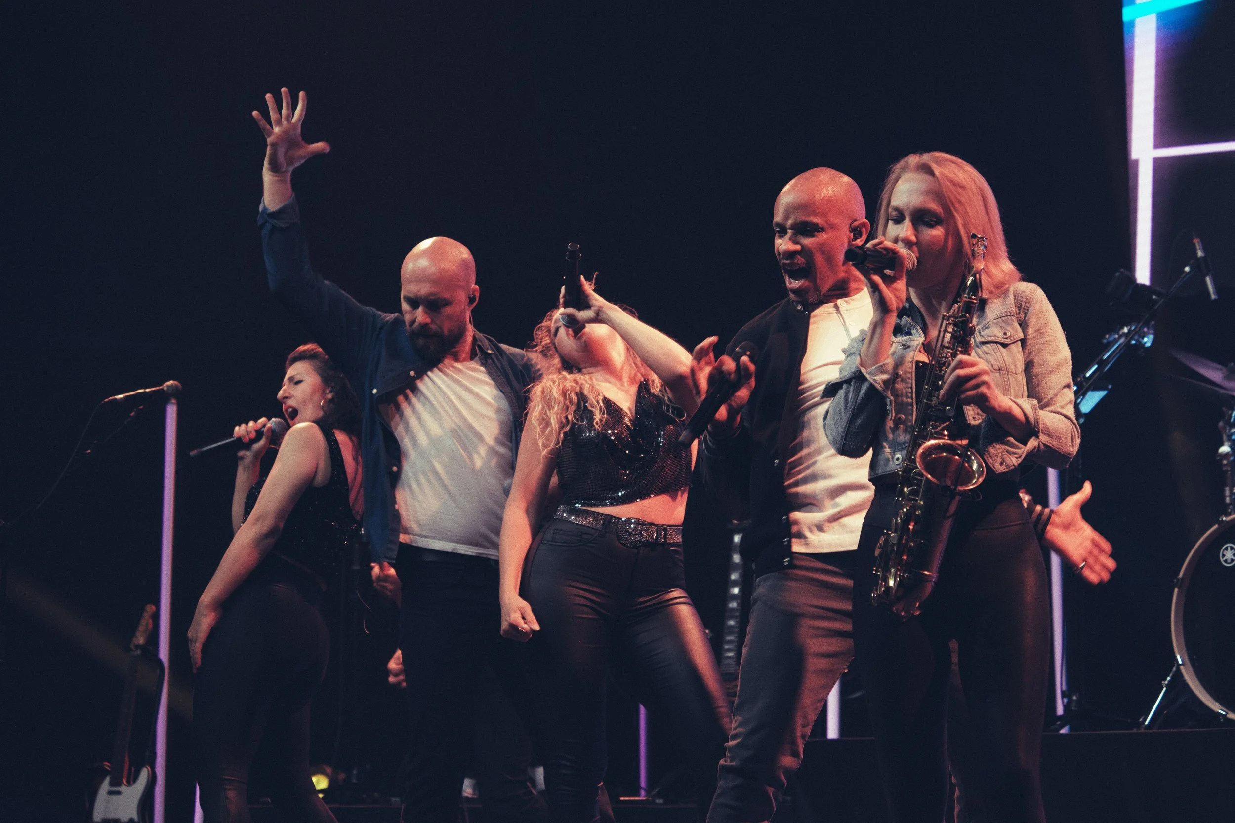 Auf einer Bühne performen fünf Musiker mit Mikrofonen, Saxophon und Gitarre, beleuchtet mit bunten Lichtern.