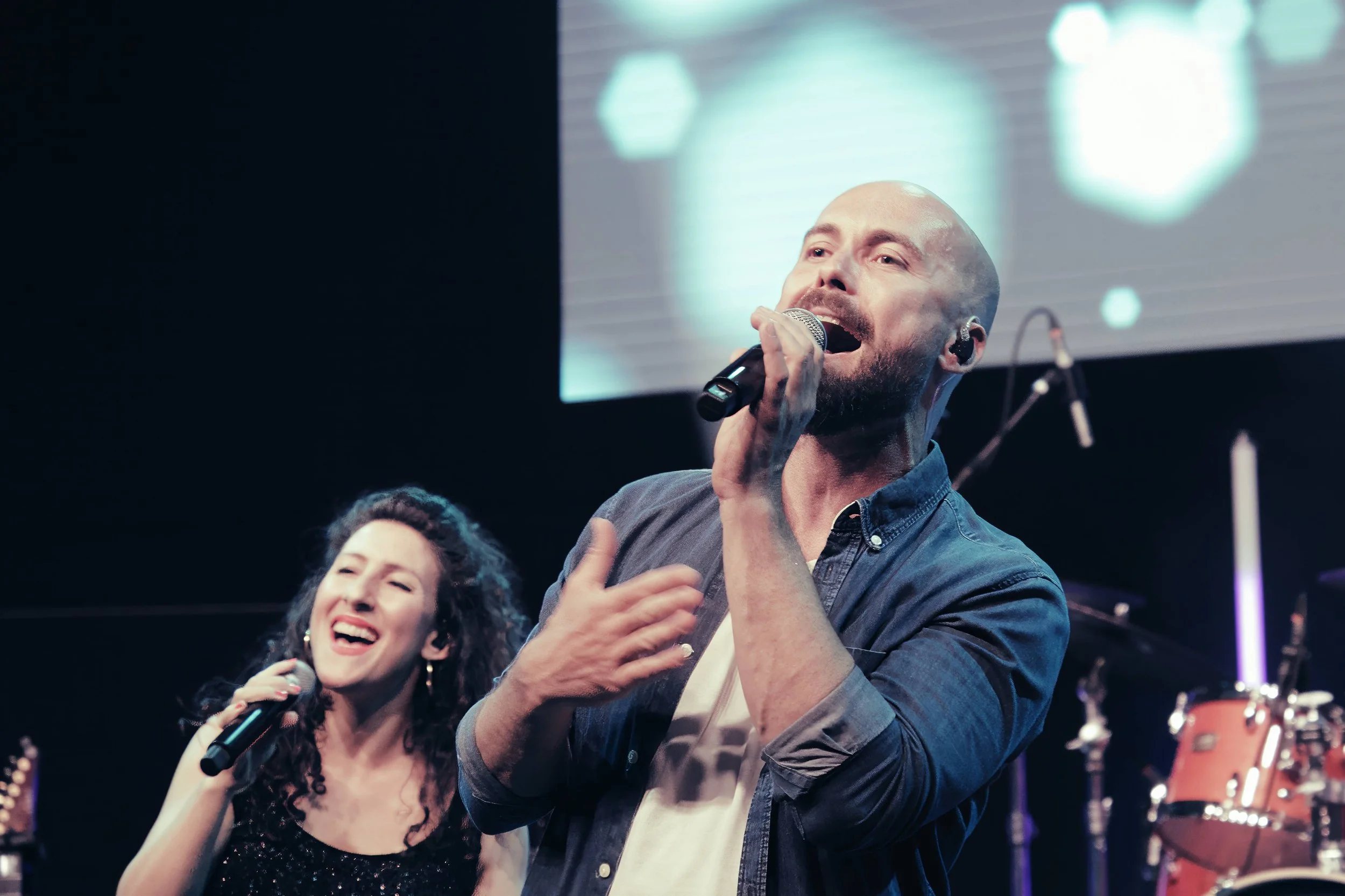 Zwei Sänger auf einer Bühne beim Singen, ein Mann mit deutschem Hemd, eine Frau mit schwarzen Locken, im Hintergrund musikalische Instrumente und ein Bildschirm mit bläulichen Mustern.