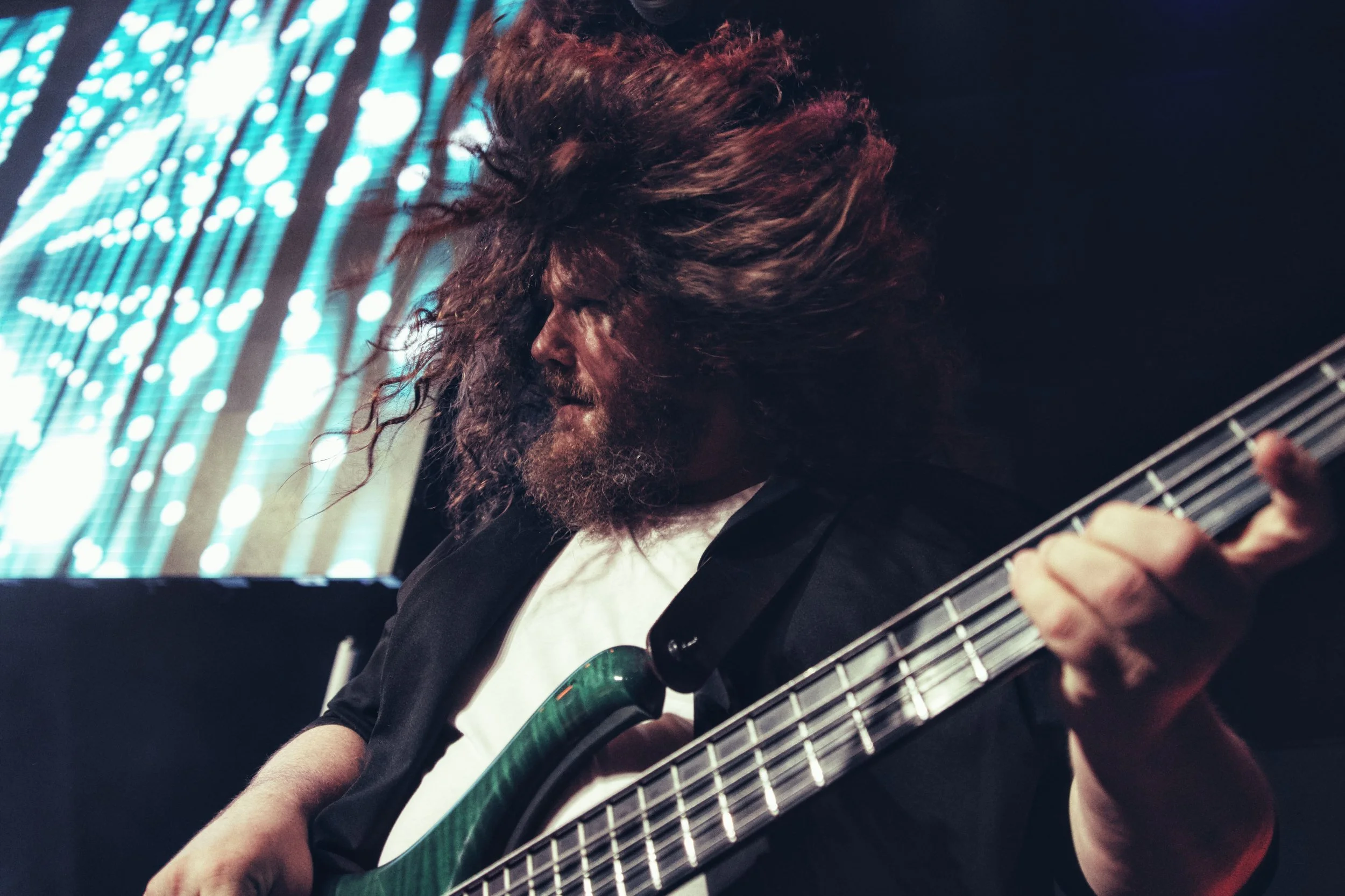 Ein Mann spielt Gitarre auf einer Bühne mit bunten Lichtern im Hintergrund, hat lange, lockige Haare und trägt dunkle Kleidung.