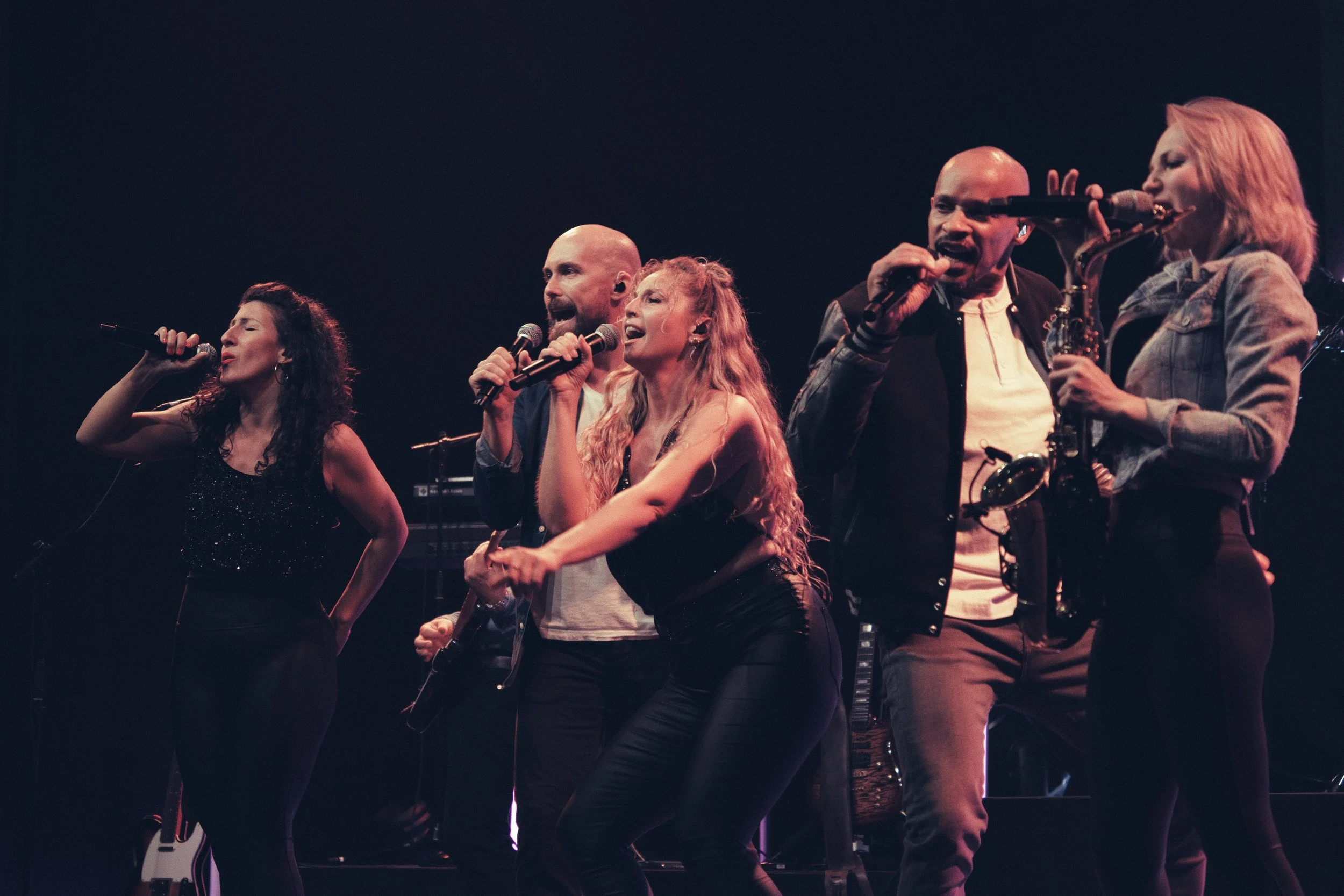Fünf Musiker beim Singen und Spielen auf der Bühne, darunter drei Frauen und zwei Männer, mit dunklem Hintergrund und Bühnenbeleuchtung.
