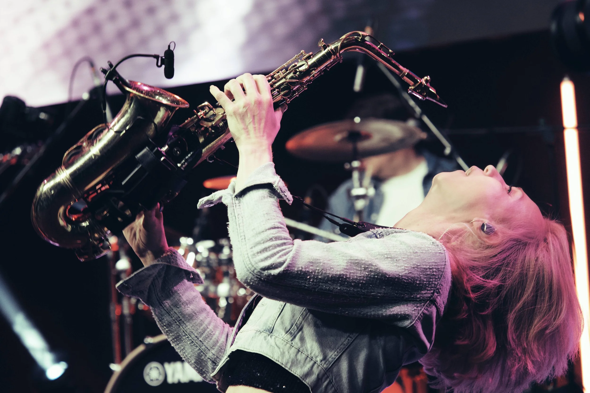 Eine Frau spielt auf einer goldfarbenen Saxophon während eines Live-Musikauftritts. Im Hintergrund sind eine Trommel und ein Schlagzeug zu sehen.