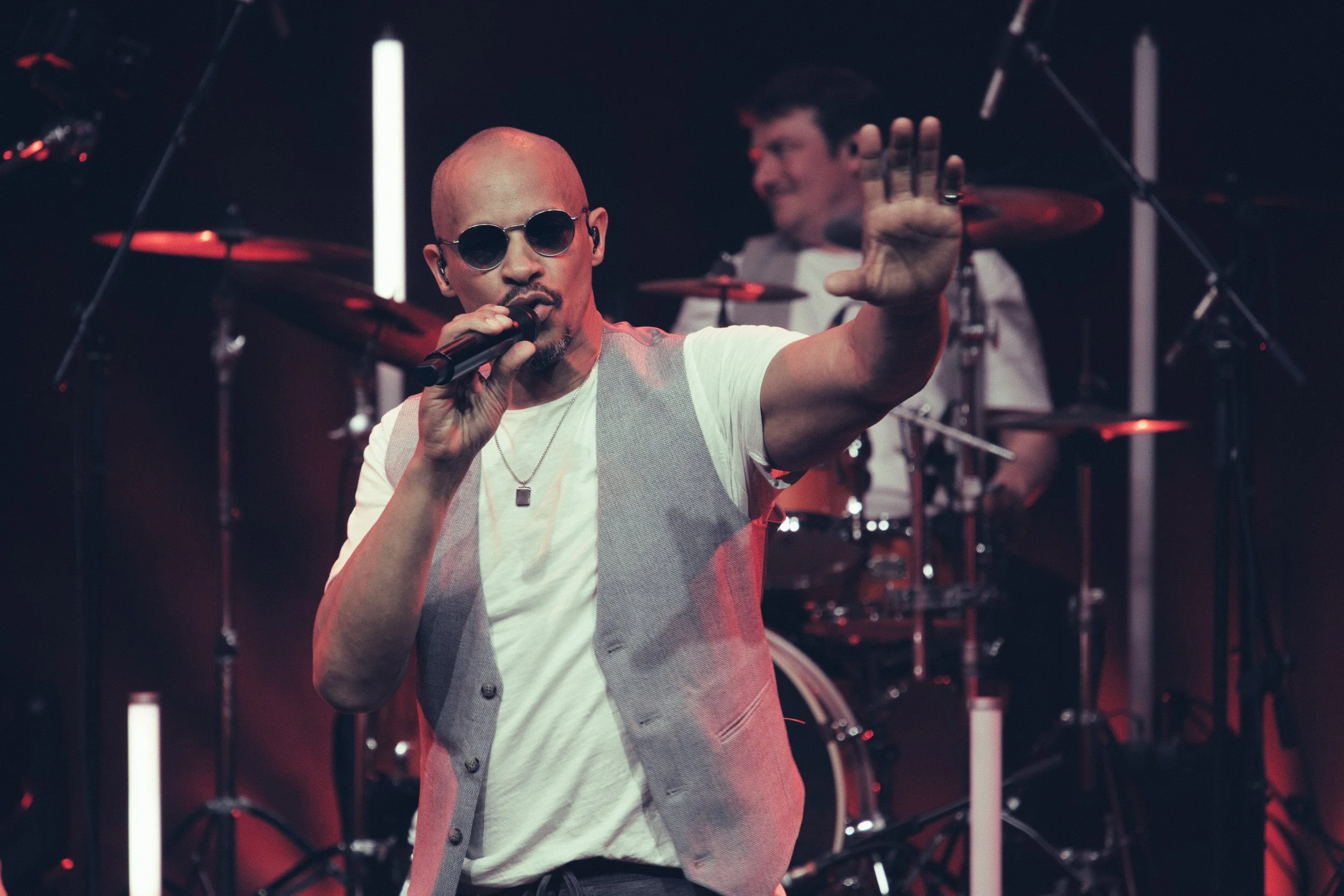 Ein Sänger mit Sonnenbrille, weißem T-Shirt und Weste singt mit einem Mikrofon, im Hintergrund ein Schlagzeuger bei einer Live-Performance auf einer Bühne.