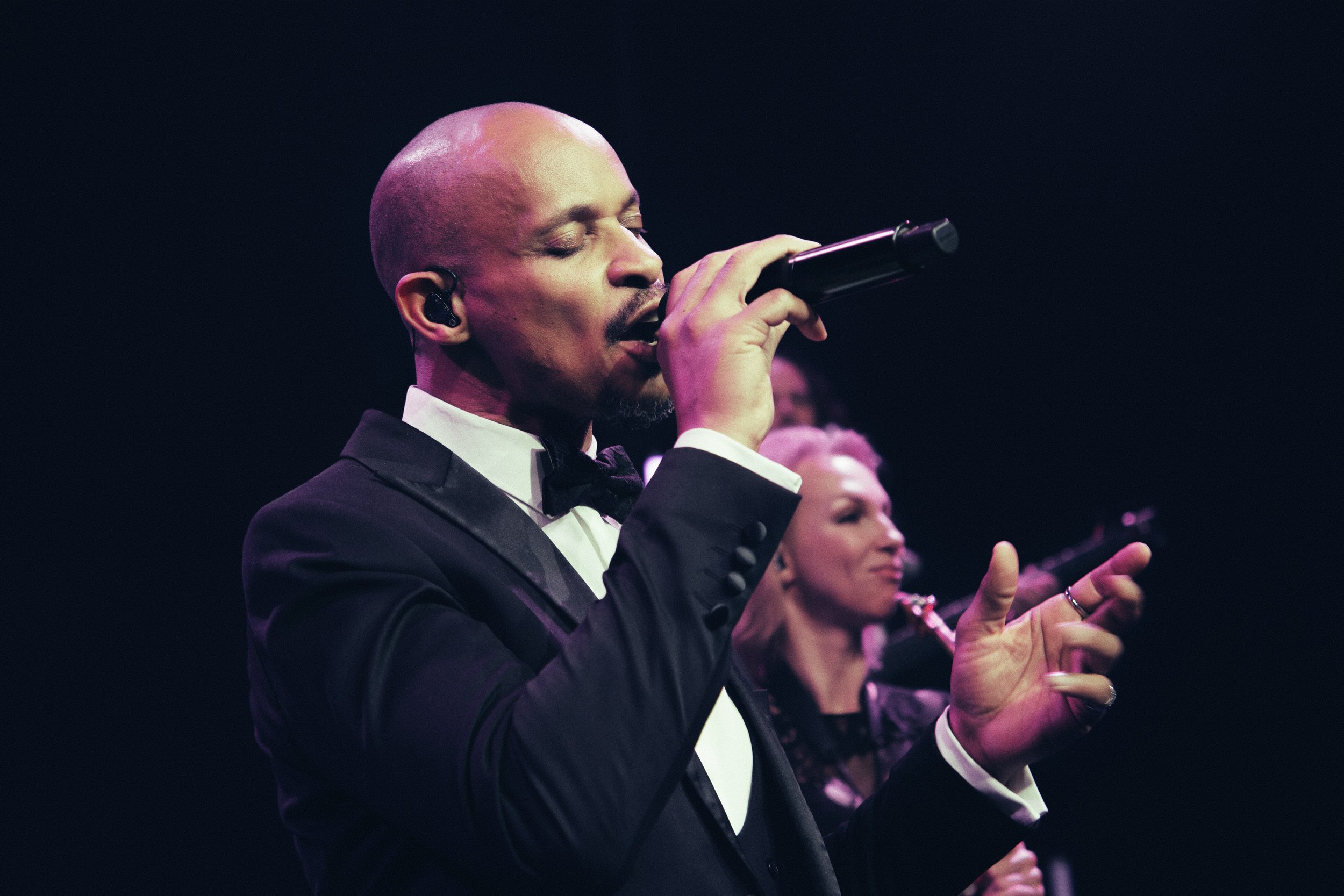 Ein Mann in einem schwarzen Anzug singt mit geschlossenen Augen in ein Mikrofon, während im Hintergrund eine Frau ebenfalls singt.