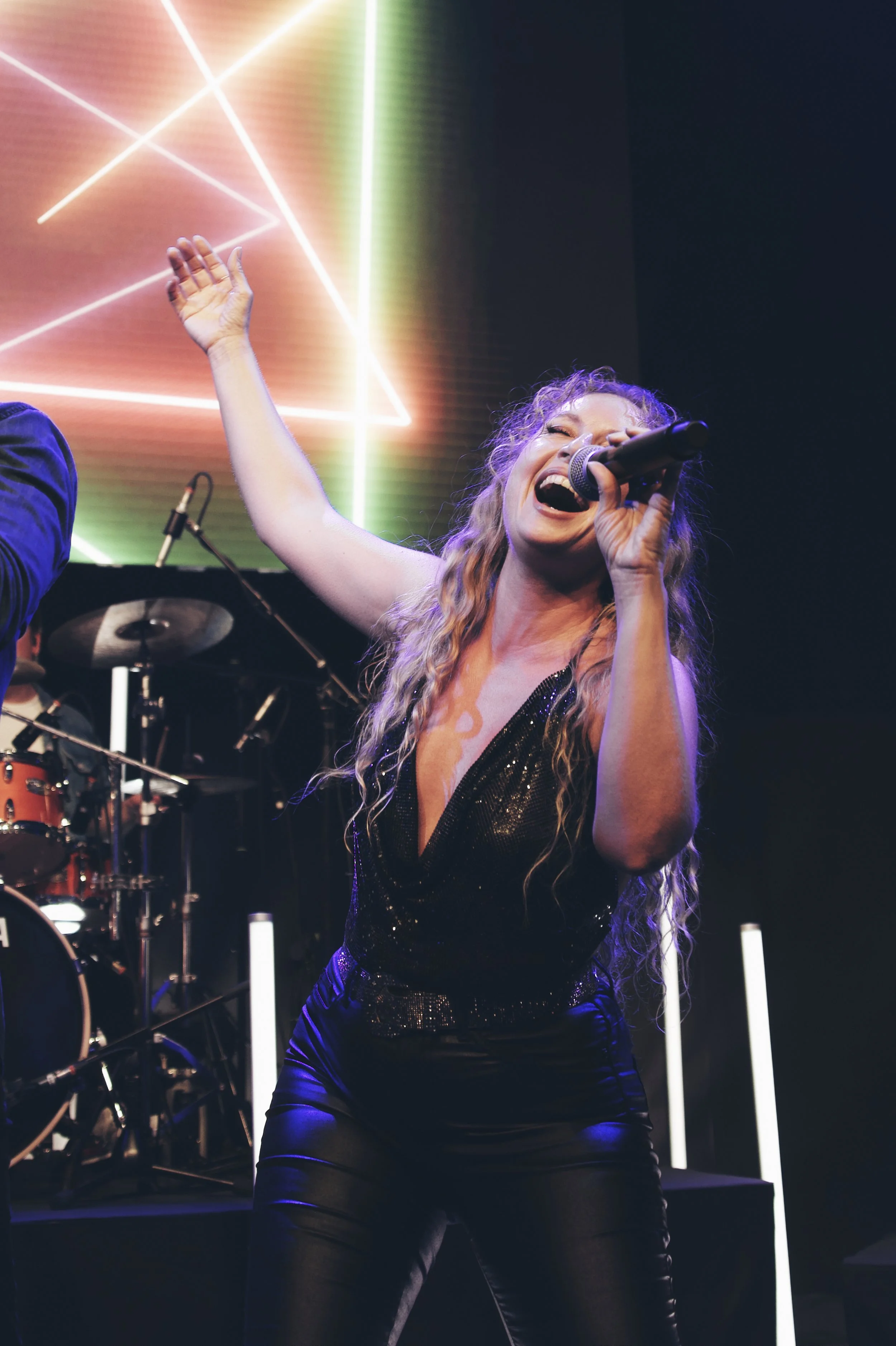 Eine Frau singt bei einem Konzert, trägt ein schwarzes, funkelndes Outfit, hat lange, lockige Haare und ist in einer dynamischen Pose mit einem Mikrofon in der Hand. Im Hintergrund sind Neonlichter und ein Schlagzeug sichtbar.