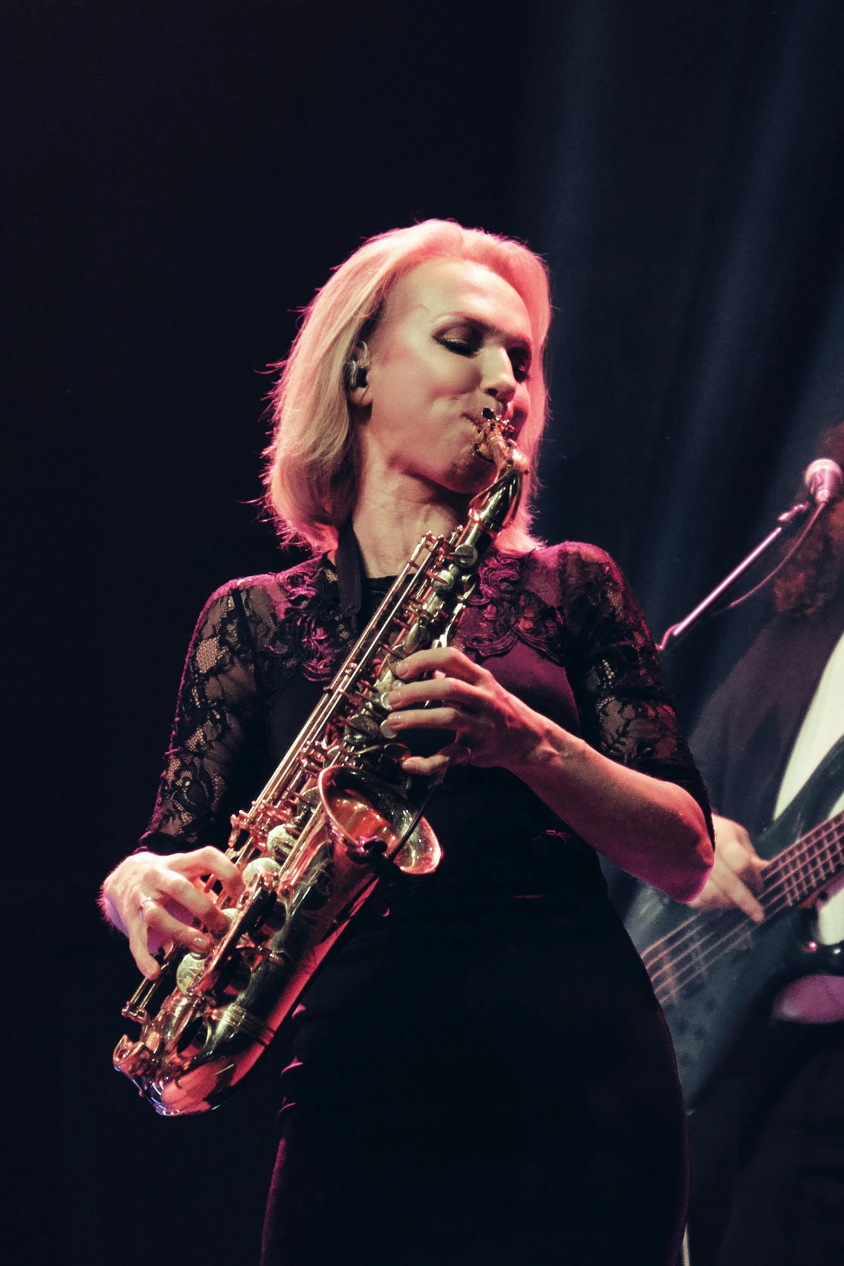 Eine Frau spielt Saxophon auf einer Bühne, im Hintergrund ist eine Gitarre zu sehen.