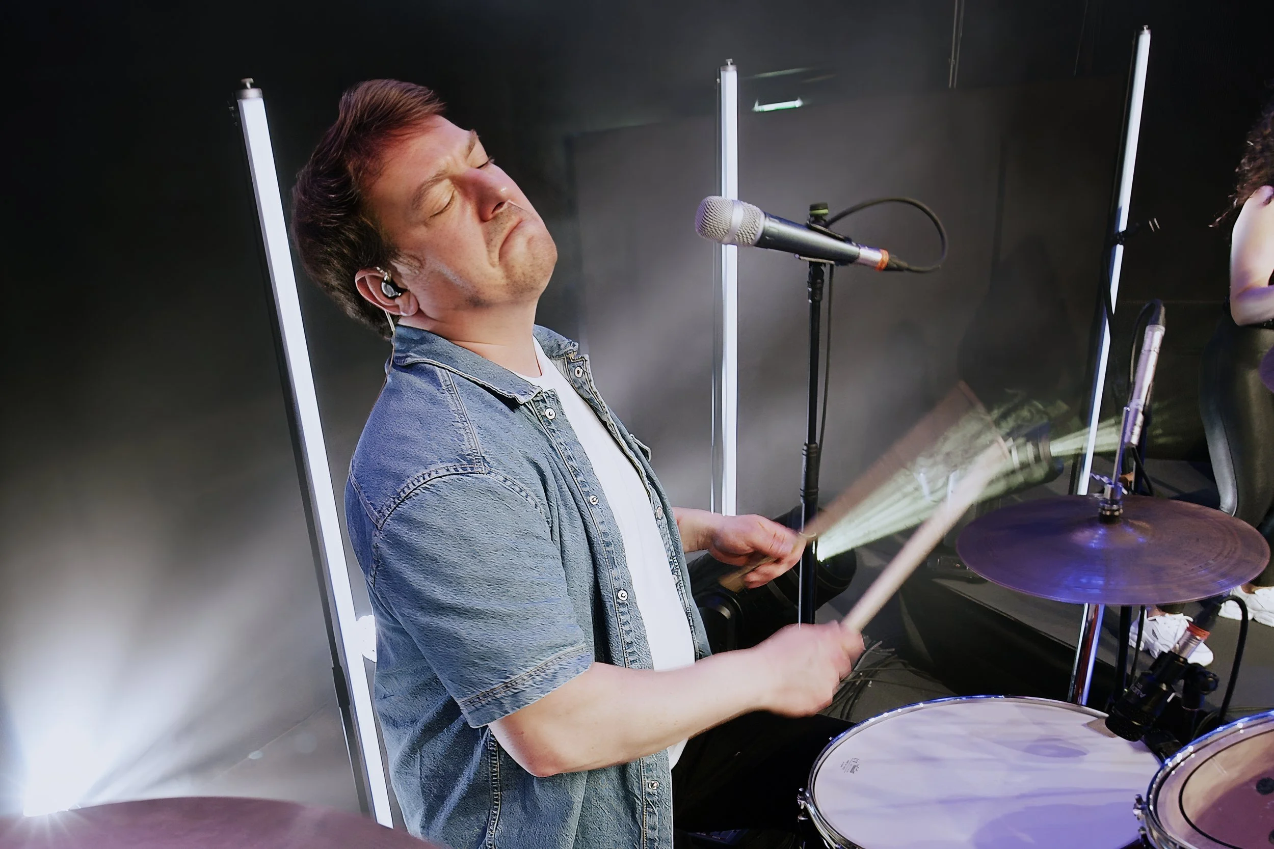 Mann spielt Schlagzeug auf einer Bühne, trägt ein Jeanshemd und hat die Augen geschlossen.