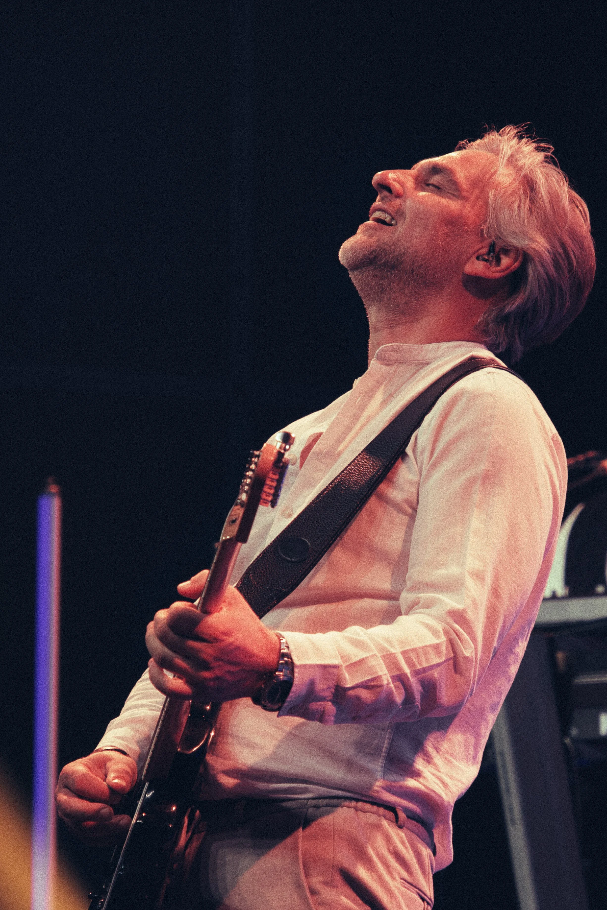 Ein älterer Mann mit grauem, lockigem Haar spielt Gitarre auf einer Bühne, sein Kopf ist nach hinten geneigt, mit einem Ausdruck von Freude oder Leidenschaft.