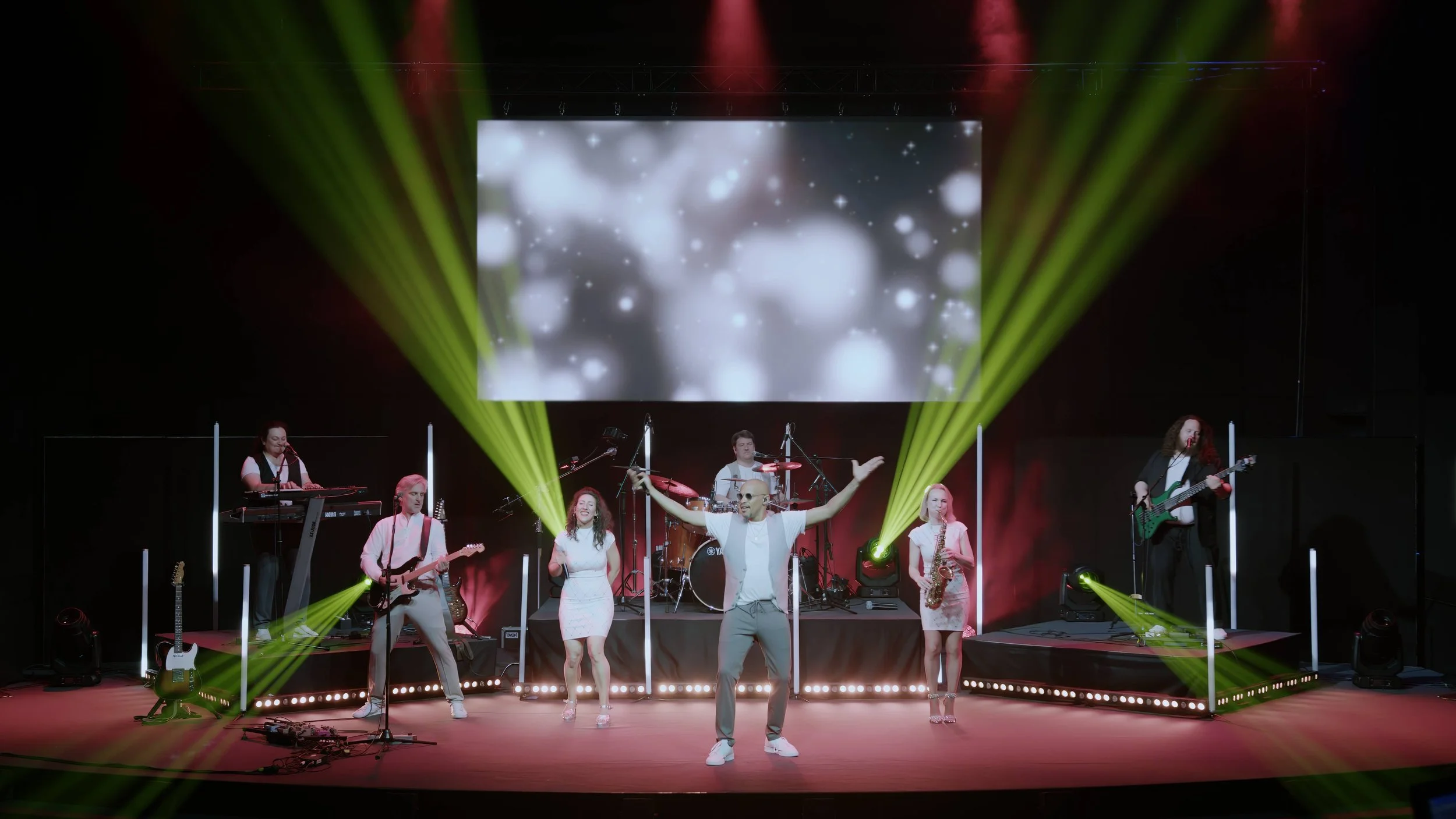 Musicalband mit sechs Mitgliedern auf einer Bühne, mit einem großen Bildschirm im Hintergrund, farbige Scheinwerfer und bunte Lichter, beim Auftritt.