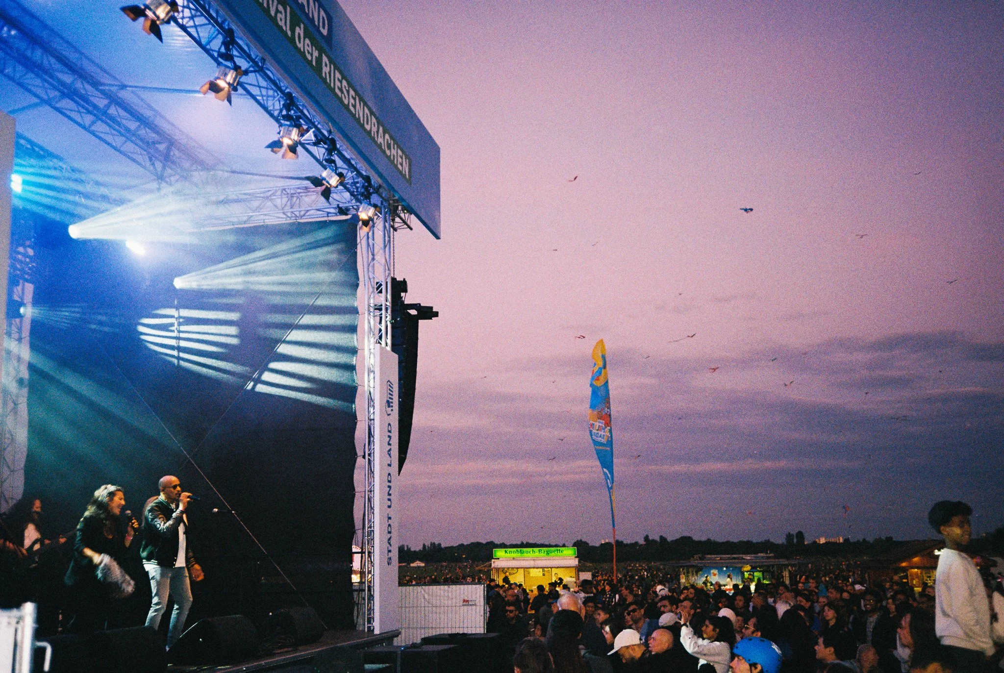 Ein Bühnenbild bei einem Festival, mit einer großen Menschenmenge im Freien, die unter einem Abendhimmel versammelt ist. Die Bühne ist mit Lichter und einem Banner versehen, auf dem 'Stadt und Land' steht, und es gibt einen Kiosk im Hintergrund.