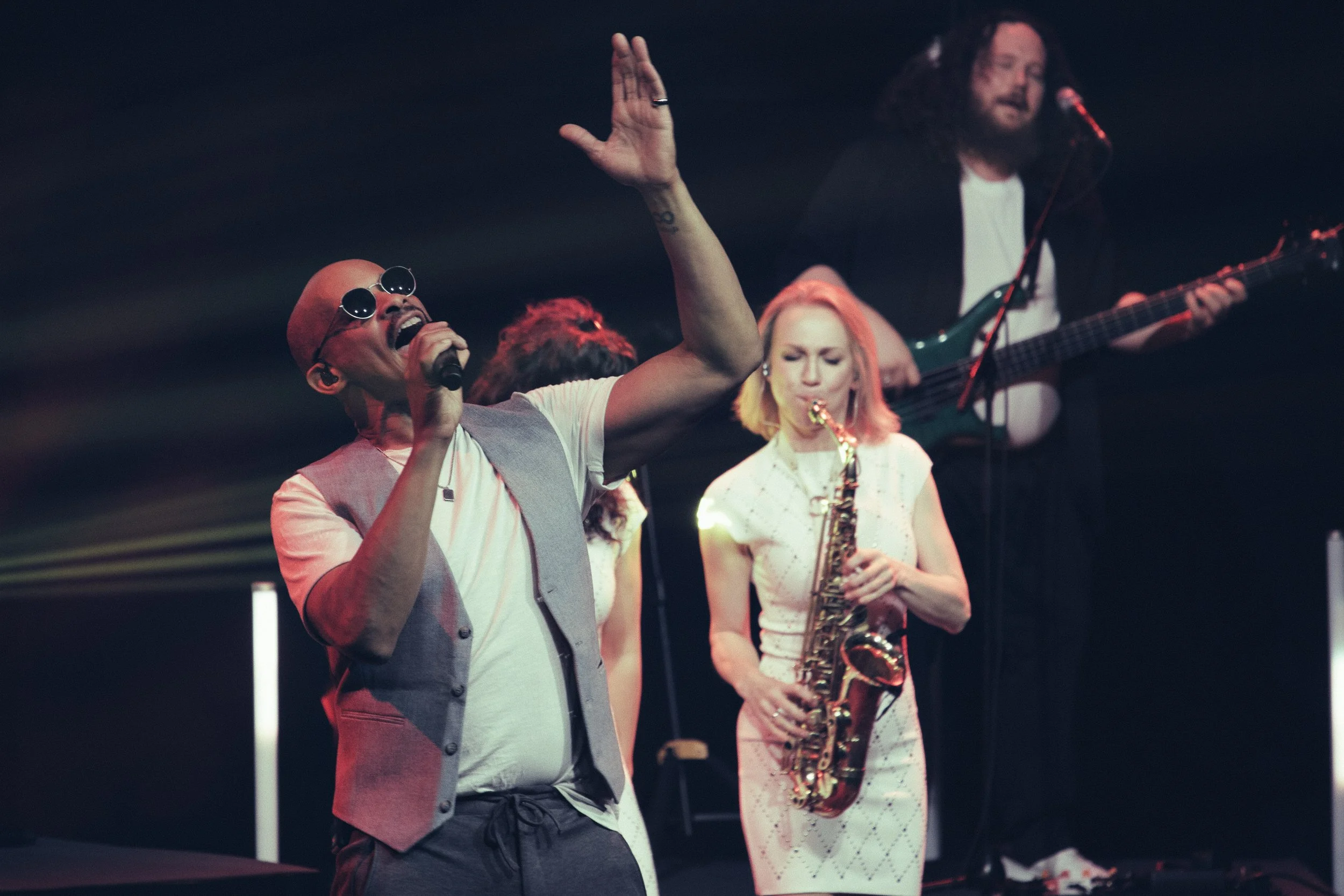 Musiker beim Live-Konzert: Sänger trägt Sonnenbrille und tanzt, Saxophonistin spielt, Gitarrist im Hintergrund, dunkler Bühnenhintergrund.