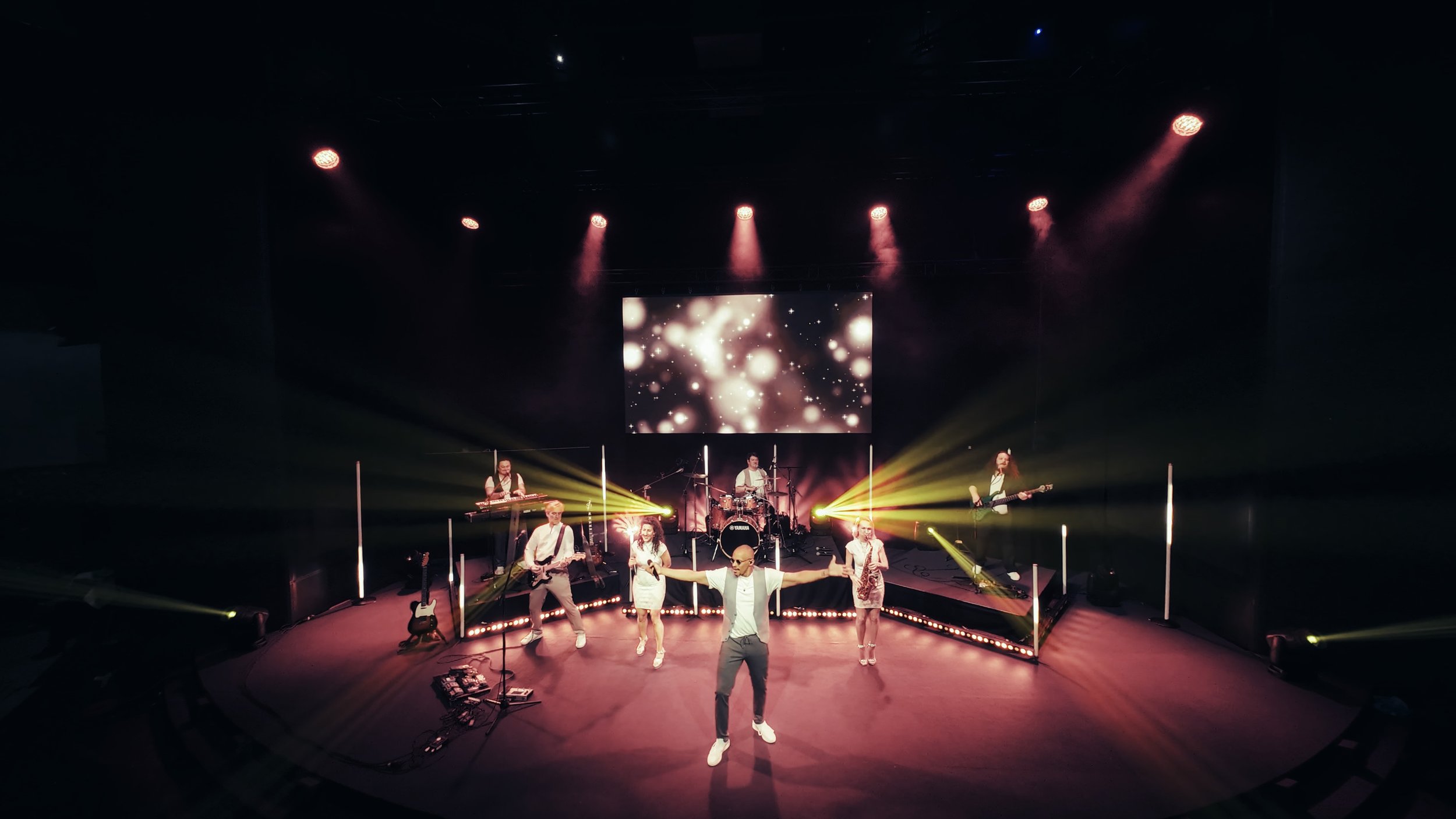 Eine Band spielt auf einer Bühne mit einer Gruppe von Sängern und Musikern, die mit Lichtern und einem großen Bildschirm im Hintergrund eine Bühnenshow präsentieren.