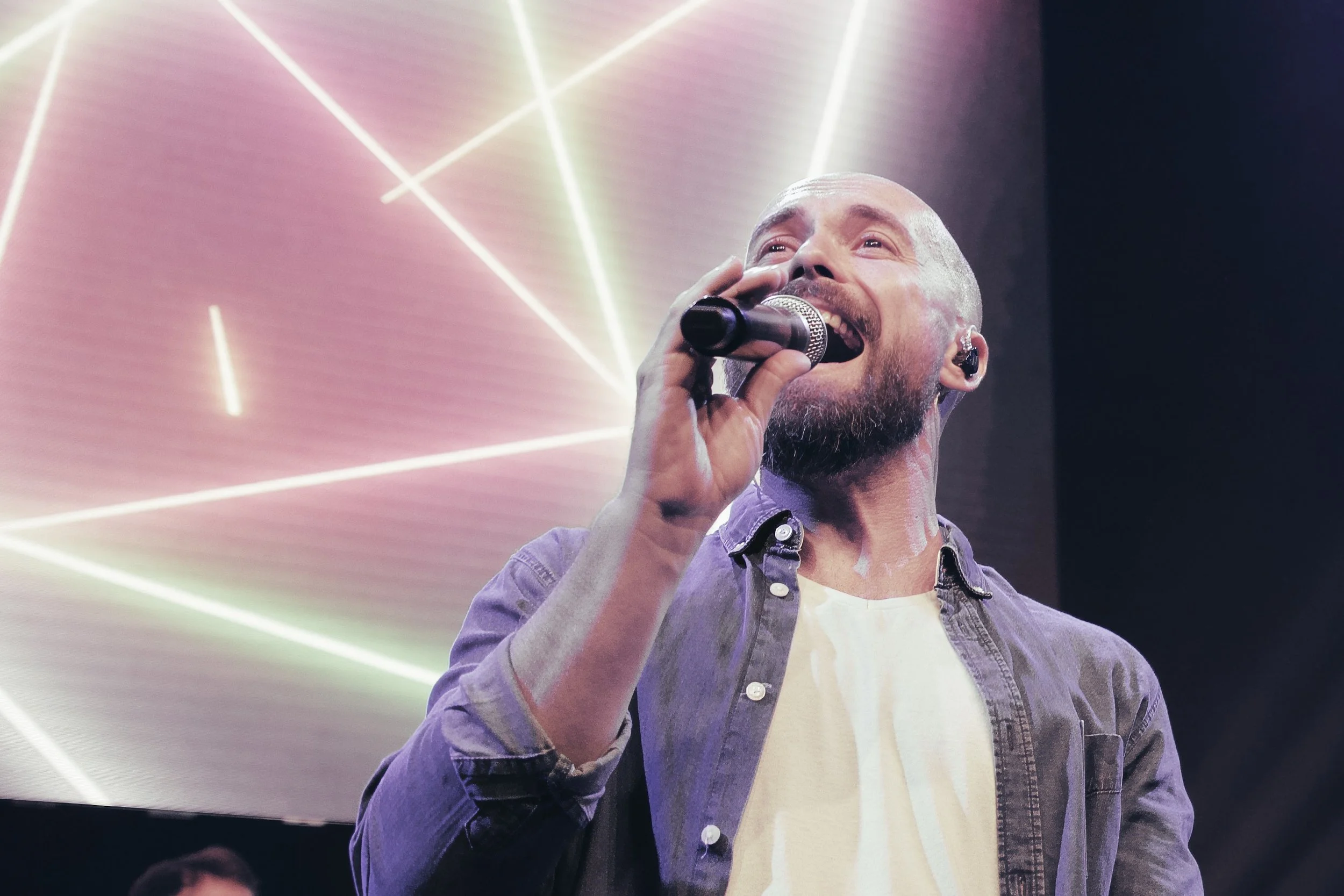 Ein Mann singt mit einem Mikrofon auf einer Bühne, im Hintergrund sind bunte Lichteffekte.