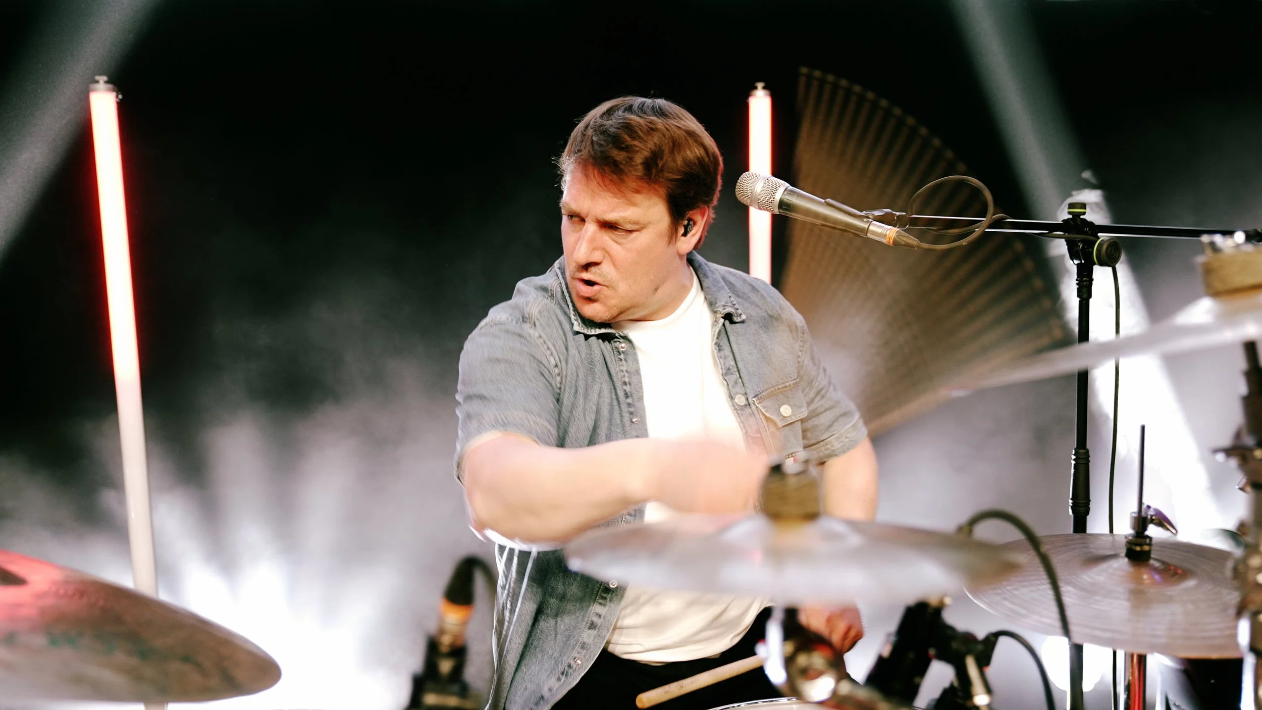 Mann spielt Schlagzeug auf einer Bühne mit Mikrofon und roter Beleuchtung.