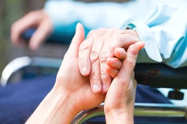 A person holding an elderly person's hand, with the elderly person lying in a hospital bed in the background.