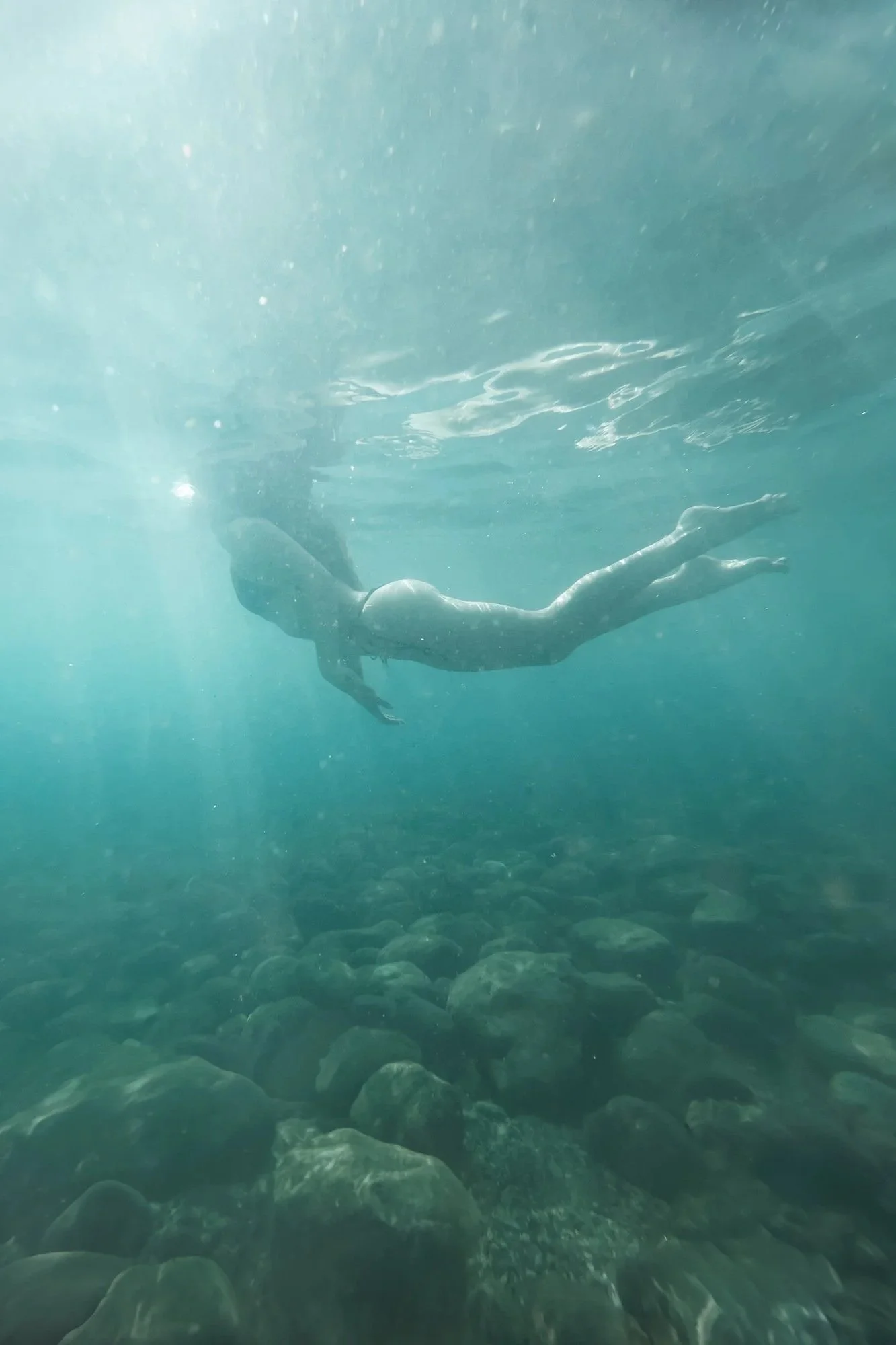 Donna che nuota sott'acqua con lo sguardo rivolto verso il basso, luce solare che attraversa l'acqua, fondo roccioso visibile.