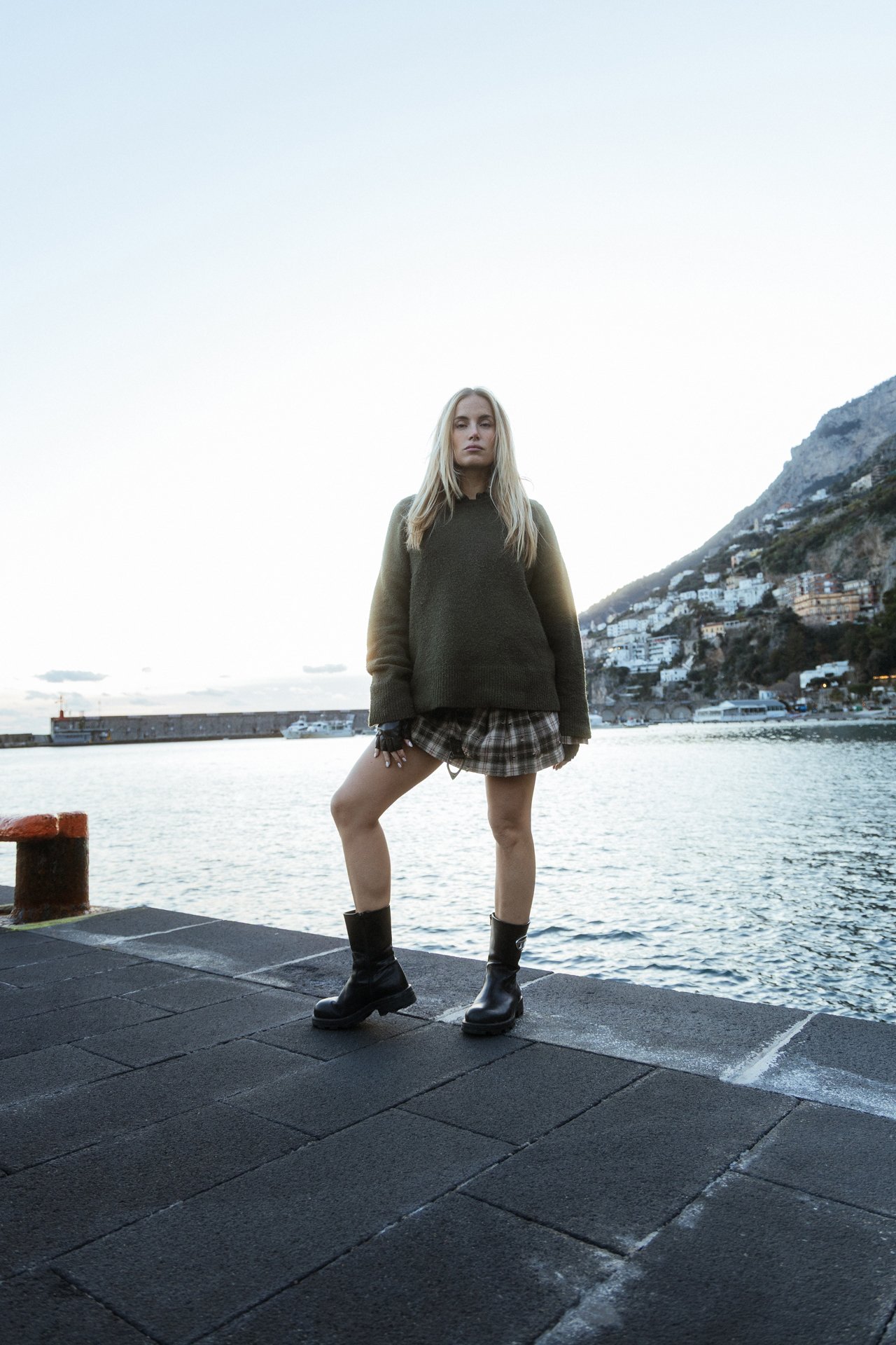 Ragazza che posa sul molo vicino a un mare con colline e case sullo sfondo, indossa un maglione verde, una minigonna scozzese e stivali neri.