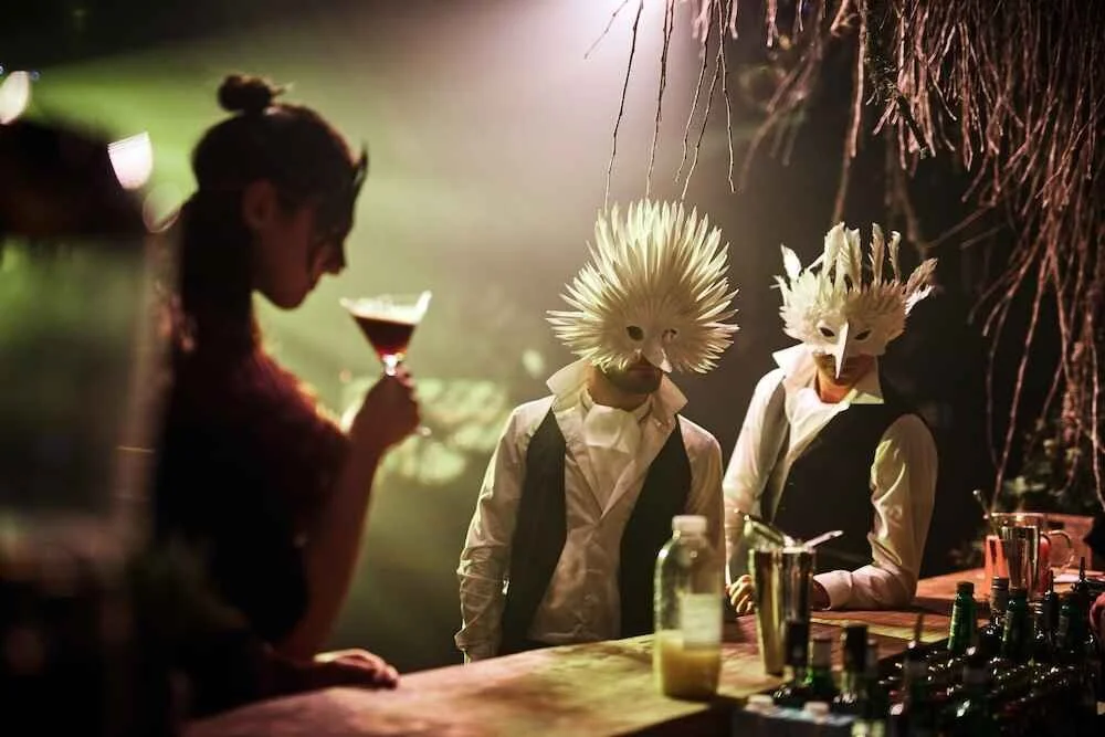 People wearing bird masks and suits at a bar, with one woman holding a drink.