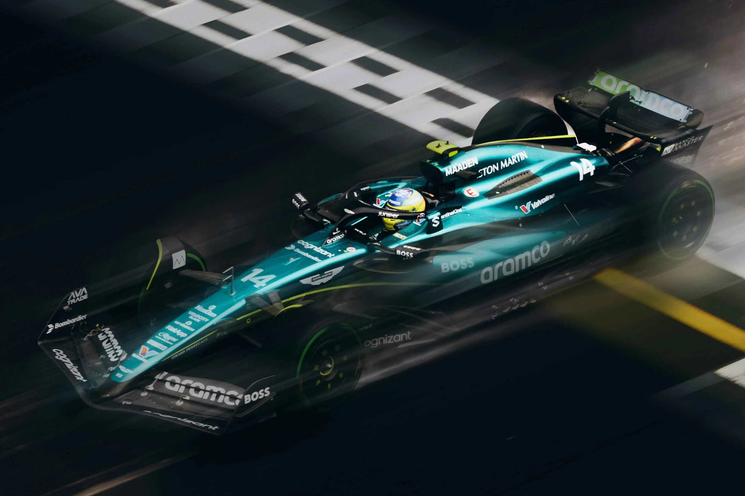 A blue Formula 2 race car speeding on a dark race track at night with motion blur, featuring the number 14 and various sponsor logos.