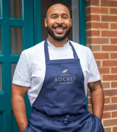 Smiling man in a white T-shirt and navy apron standing outside in front of a brick wall and blue door.