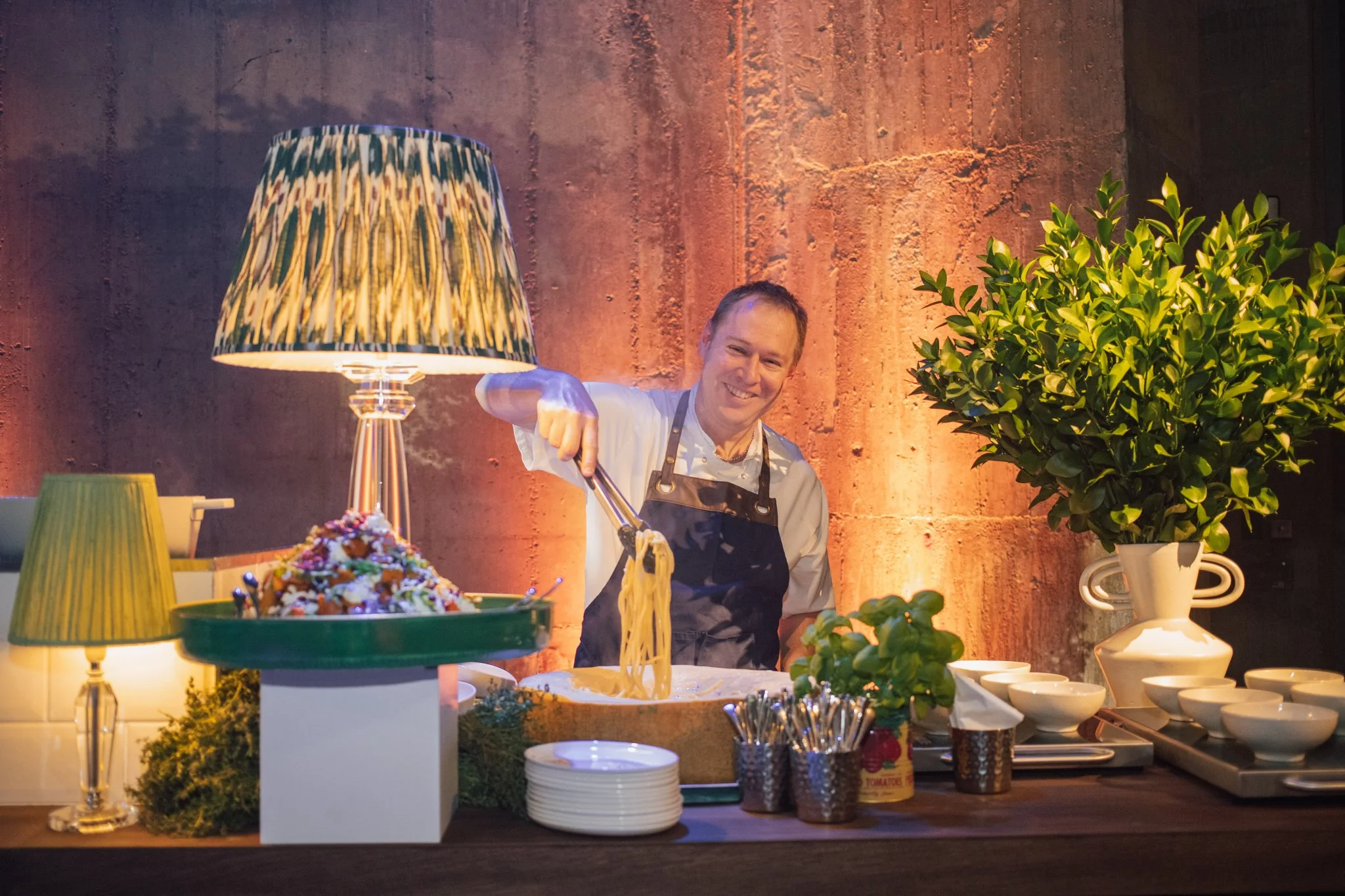 A smiling chef in a white shirt and apron preparing pasta in a dining area with decorative lamps, bowls, and a large potted plant.