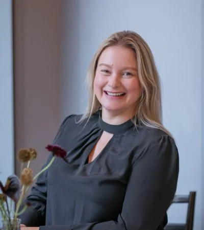 A smiling woman with blonde hair sitting at a table with flowers in a vase.