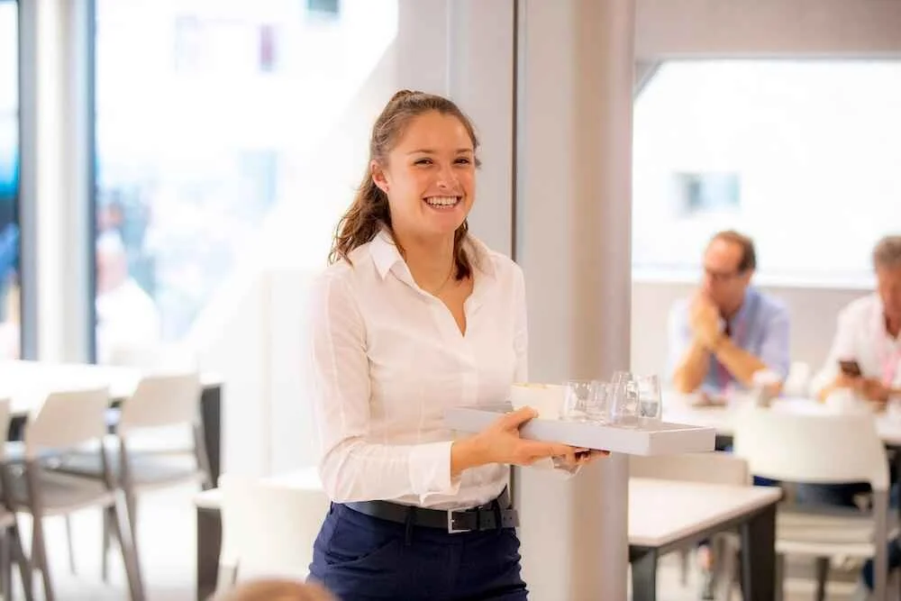 A smiling waitress holding a tray with glasses and a small plate in a bright, modern restaurant.