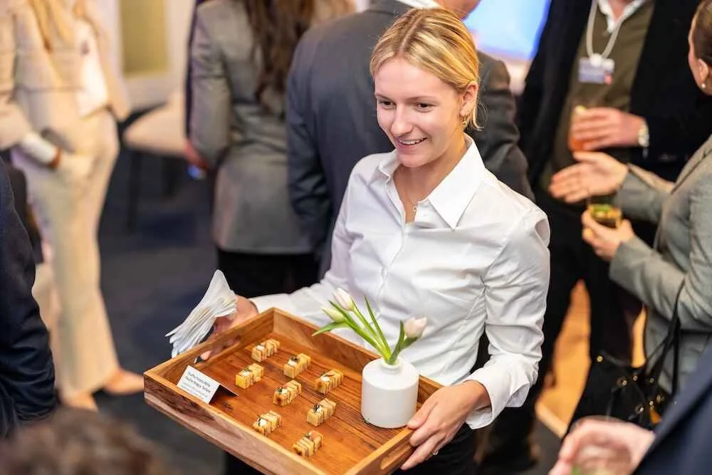 Woman serving small desserts on a wooden tray at a social event.