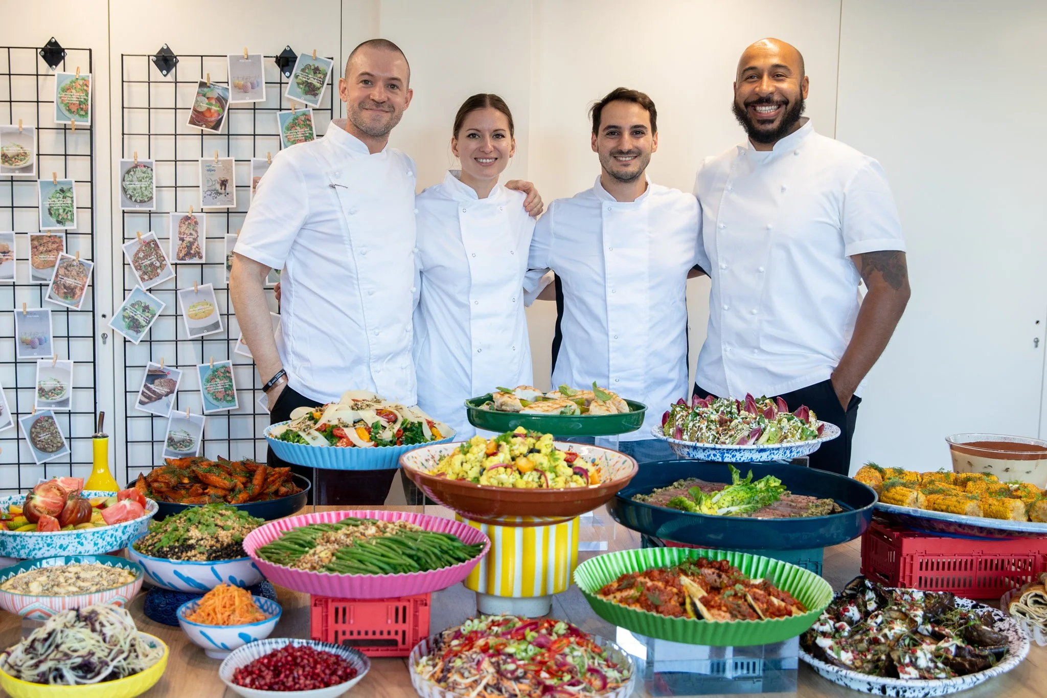 Four chefs in white uniforms standing behind a table filled with various colorful salads and dishes, with a photo display of food images in the background.