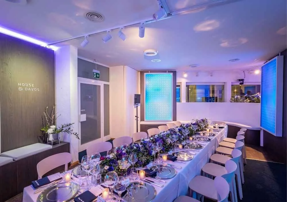 Elegant banquet room set for a formal dinner with a long table, white tablecloth, floral centerpiece, candles, wine glasses, and neatly arranged place settings, illuminated by purple and blue lighting.